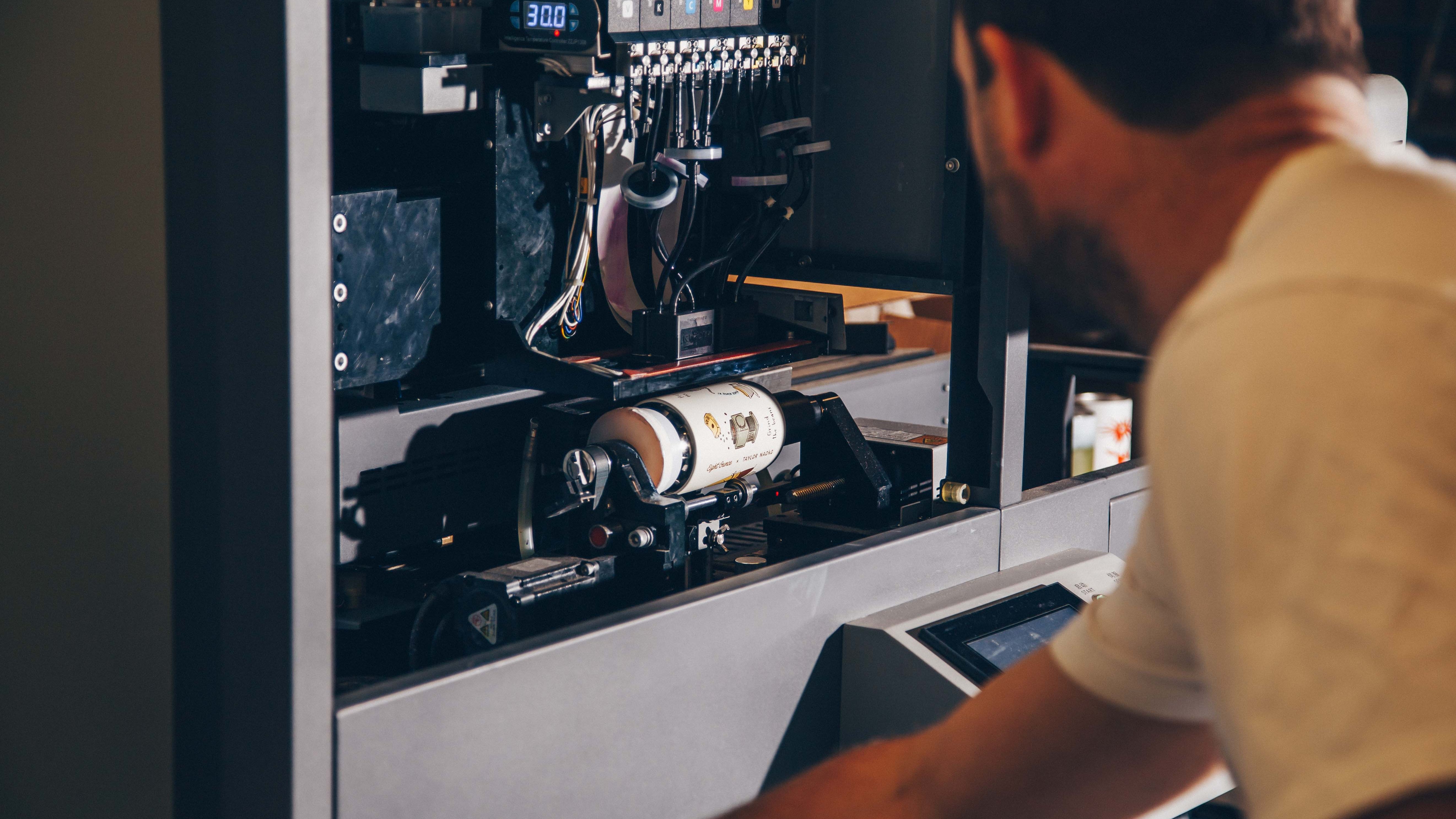 A person supervises the operation of a rotary printer, observing the design application on a bottle in real time.
