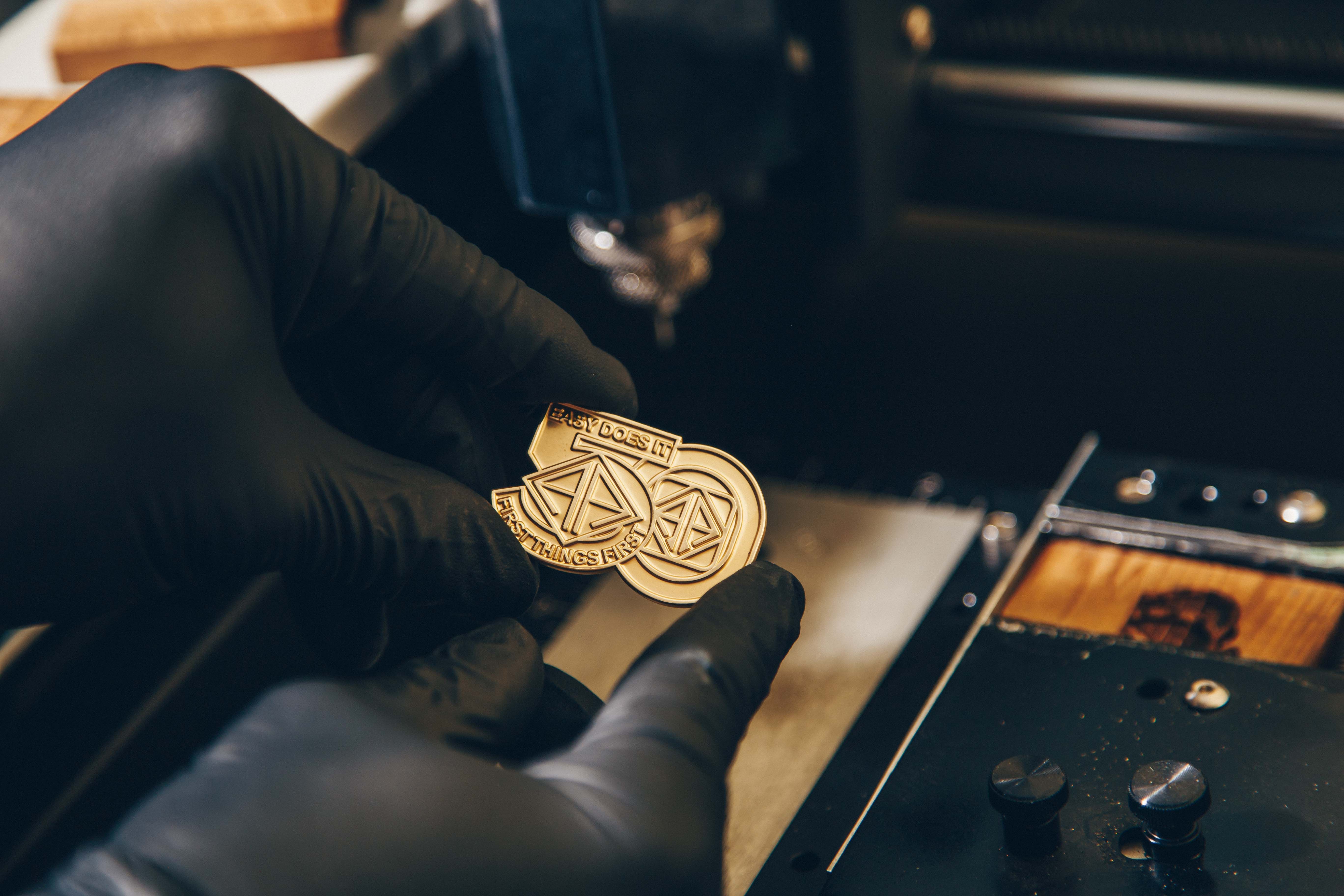 A close-up of a hand wearing a black glove holding a gold-colored metal object, possibly a coin or medallion, with intricate designs and text. The text on the object reads "EASY DOES IT" and "FIRST THINGS FIRST." The background shows a part of a machine, suggesting a workshop or manufacturing setting.