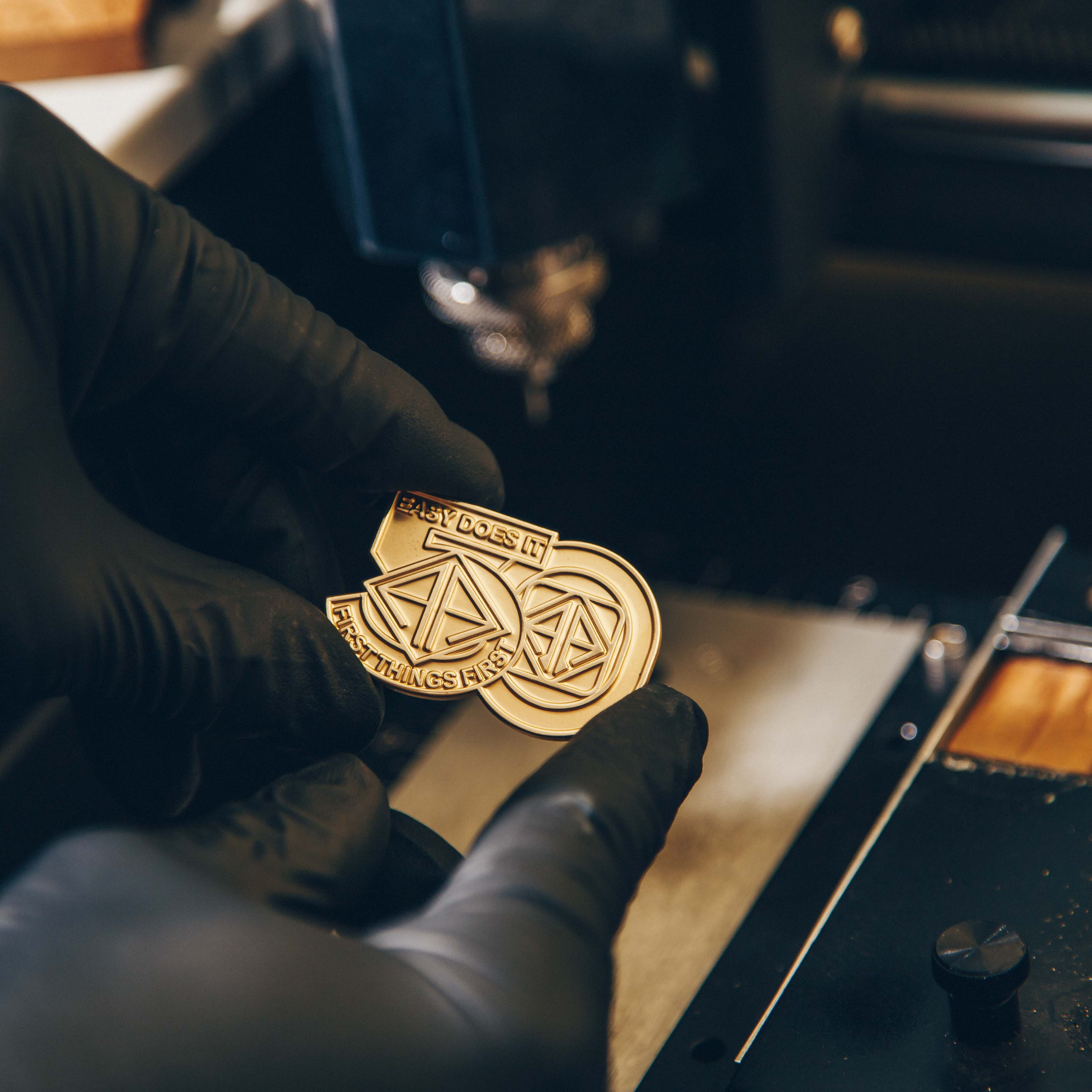 A close-up of a hand wearing a black glove holding a gold-colored metal object, possibly a coin or medallion, with intricate designs and text. The text on the object reads "EASY DOES IT" and "FIRST THINGS FIRST." The background shows a part of a machine, suggesting a workshop or manufacturing setting.