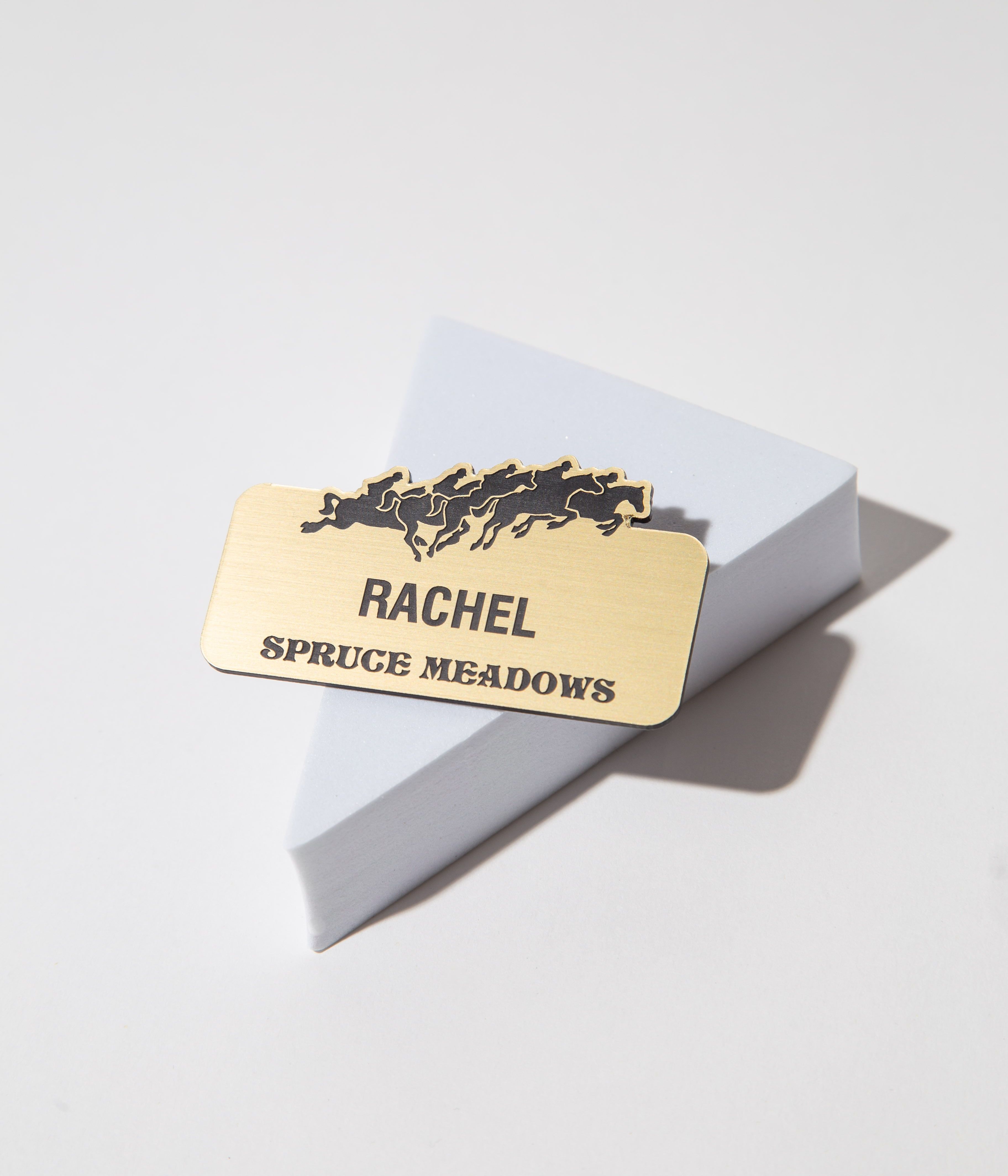 Gold name tag with horse silhouettes and text 'Rachel Spruce Meadows' on a geometric stand.