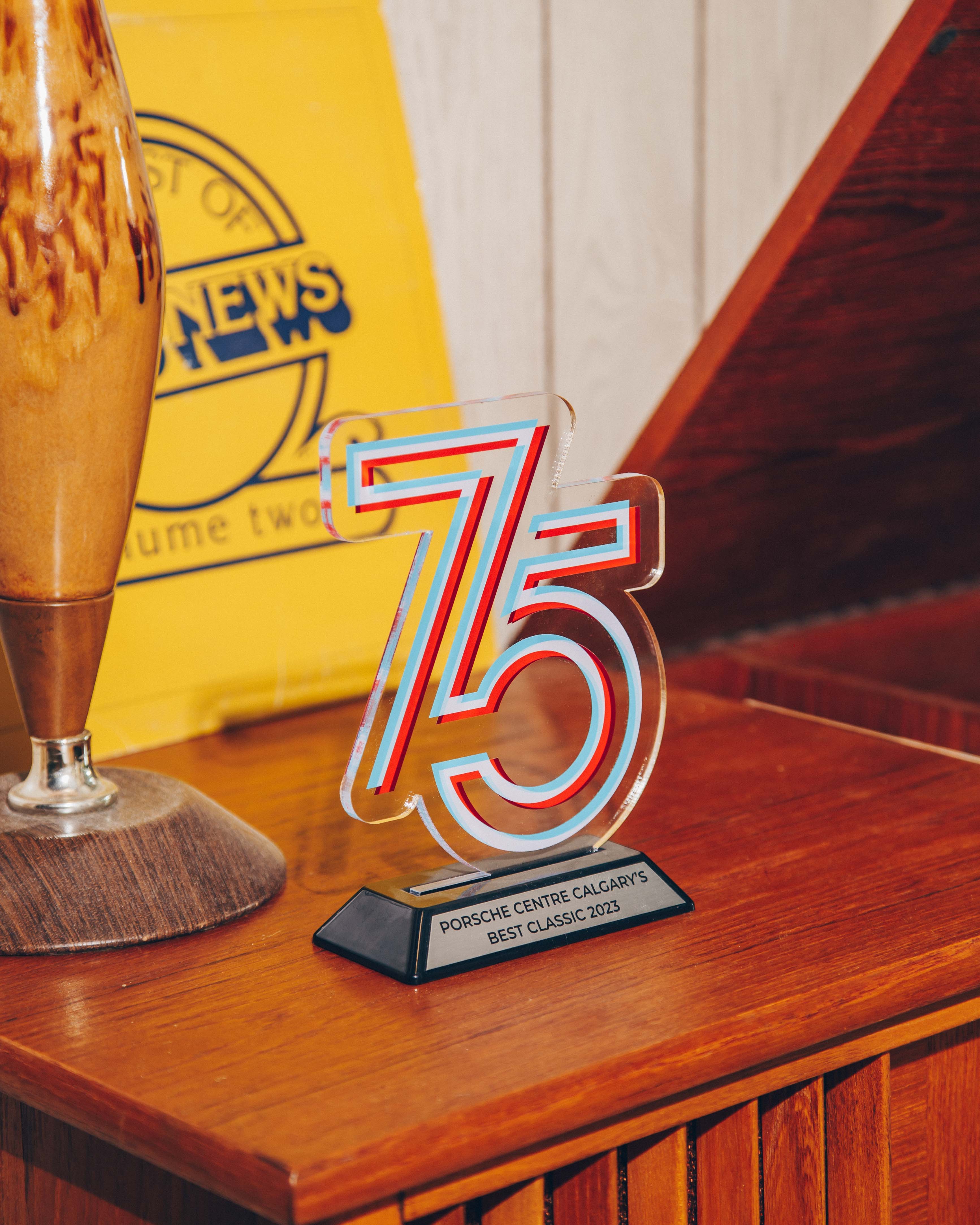A wooden sideboard on which a clear acrylic trophy with the number '75' is prominently displayed. The trophy has a modern design with red and blue accents and is mounted on a black base with a plaque that reads 'Porsche Centre Calgary's Best Classic 2023.' To the left of the trophy, there is a vintage-style lamp with a wooden base and a ceramic body. In the background, a yellow sign with blue text is partially visible, adding a retro feel to the setting. The overall composition is warm and nostalgic, with a focus on the trophy as the central subject.
