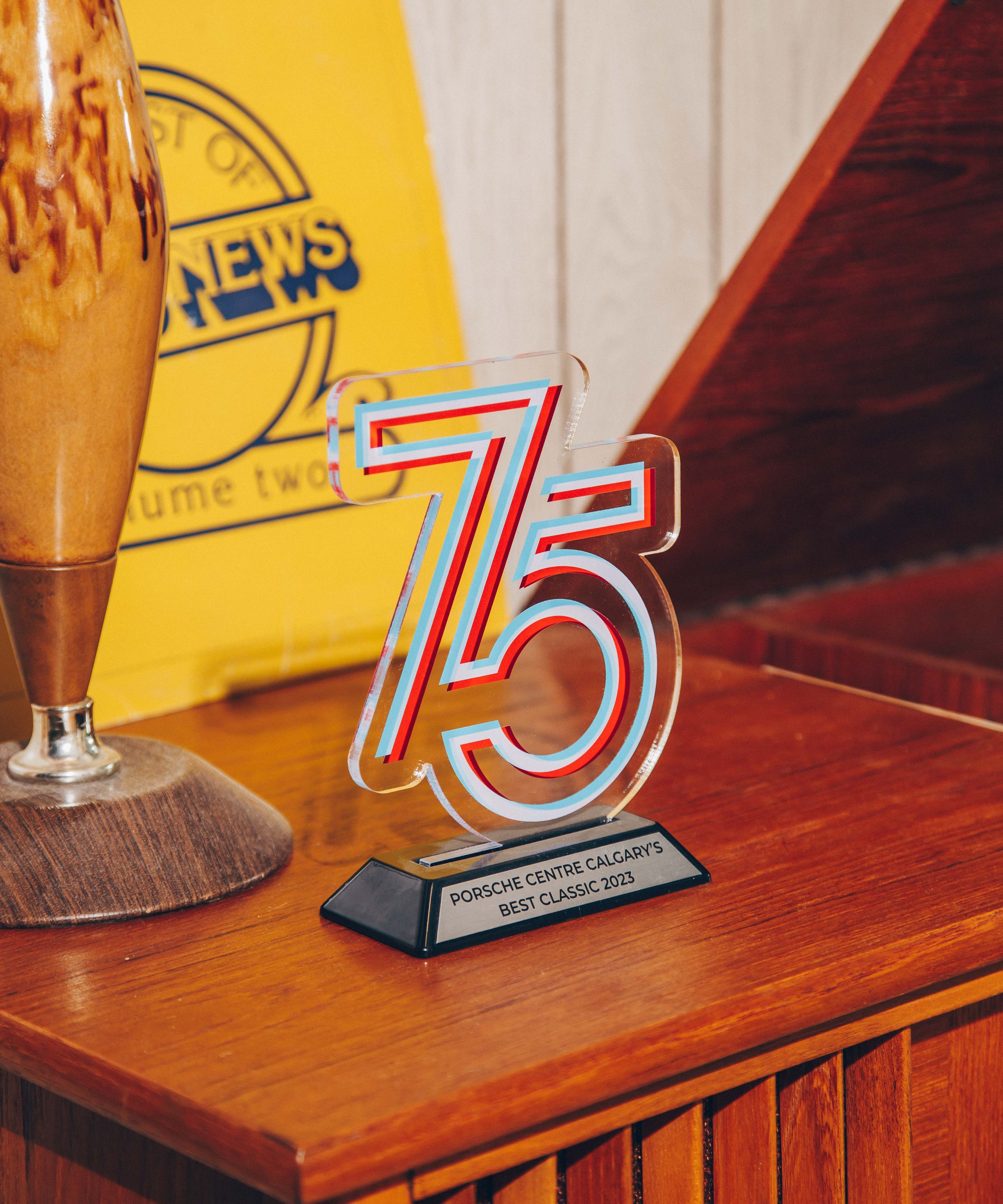 A wooden sideboard on which a clear acrylic trophy with the number '75' is prominently displayed. The trophy has a modern design with red and blue accents and is mounted on a black base with a plaque that reads 'Porsche Centre Calgary's Best Classic 2023.' To the left of the trophy, there is a vintage-style lamp with a wooden base and a ceramic body. In the background, a yellow sign with blue text is partially visible, adding a retro feel to the setting. The overall composition is warm and nostalgic, with a focus on the trophy as the central subject.