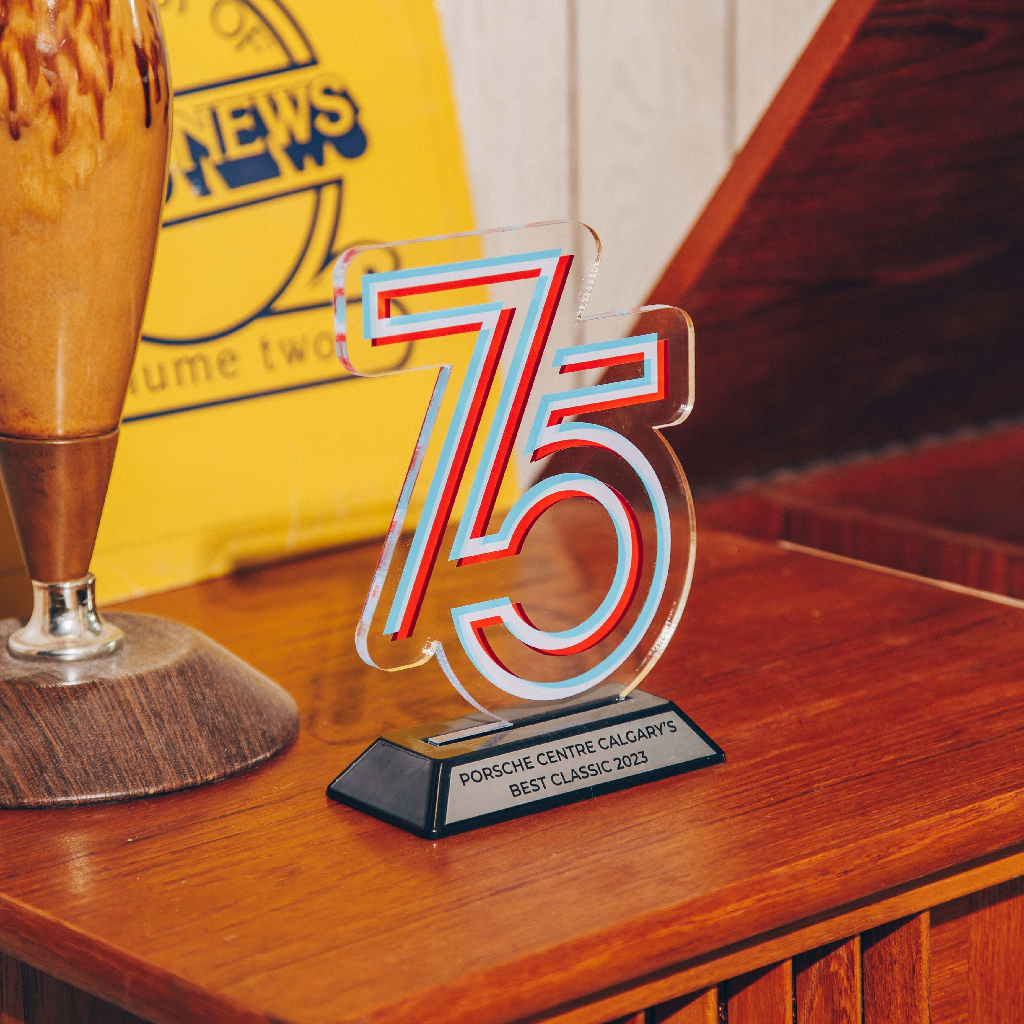 A wooden sideboard on which a clear acrylic trophy with the number '75' is prominently displayed. The trophy has a modern design with red and blue accents and is mounted on a black base with a plaque that reads 'Porsche Centre Calgary's Best Classic 2023.' To the left of the trophy, there is a vintage-style lamp with a wooden base and a ceramic body. In the background, a yellow sign with blue text is partially visible, adding a retro feel to the setting. The overall composition is warm and nostalgic, with a focus on the trophy as the central subject.