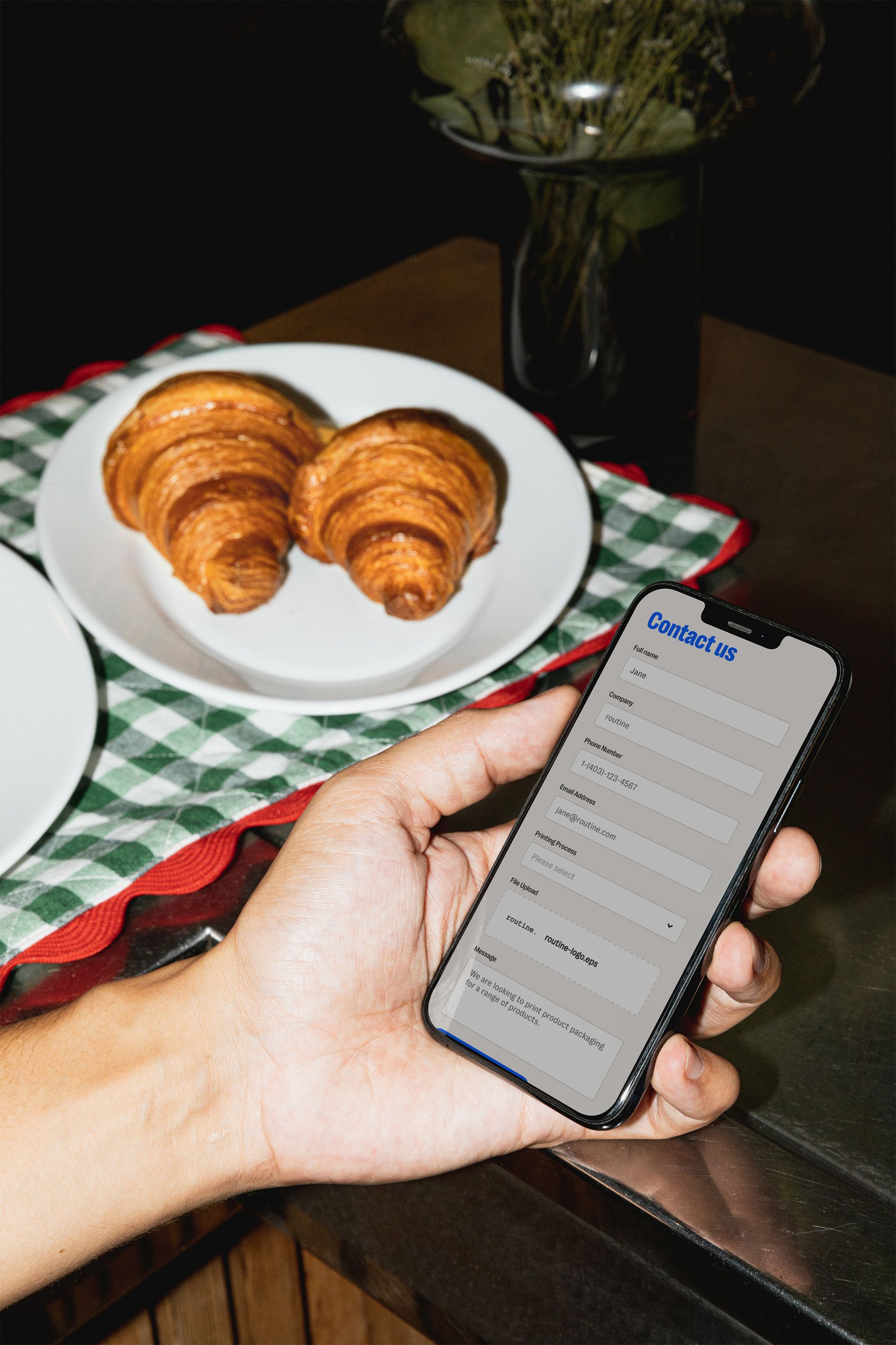 A person holding a smartphone displaying a "Contact Us" form, with fields for full name, company, phone number, email, priority, file upload, and a message section. The form appears completed with placeholder text. In the background, there is a plate of two golden-brown croissants placed on a checkered green and white cloth atop a table. A vase with green foliage is visible behind the plate, adding a casual and cozy ambiance.