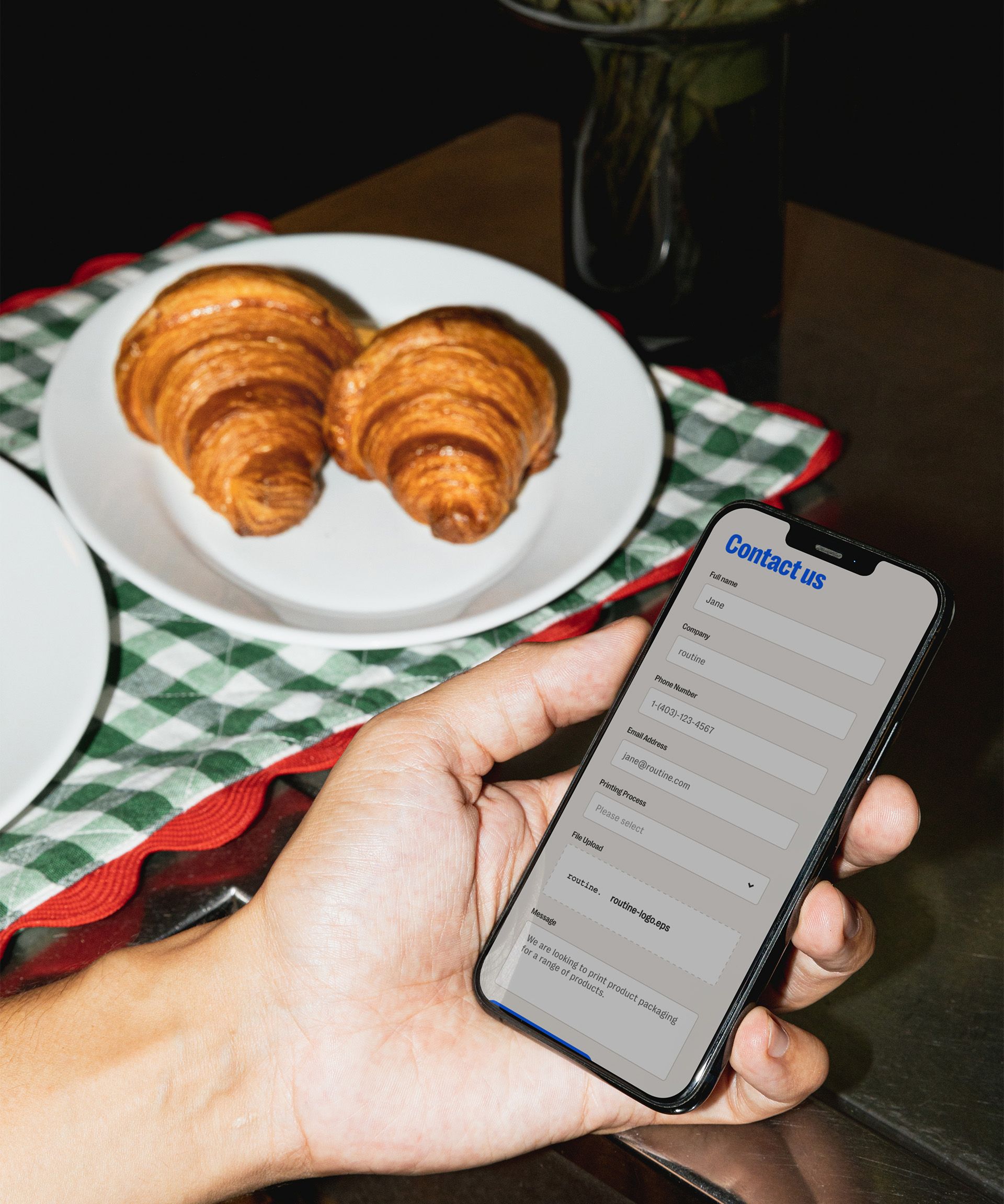 A person holding a smartphone displaying a "Contact Us" form, with fields for full name, company, phone number, email, priority, file upload, and a message section. The form appears completed with placeholder text. In the background, there is a plate of two golden-brown croissants placed on a checkered green and white cloth atop a table. A vase with green foliage is visible behind the plate, adding a casual and cozy ambiance.