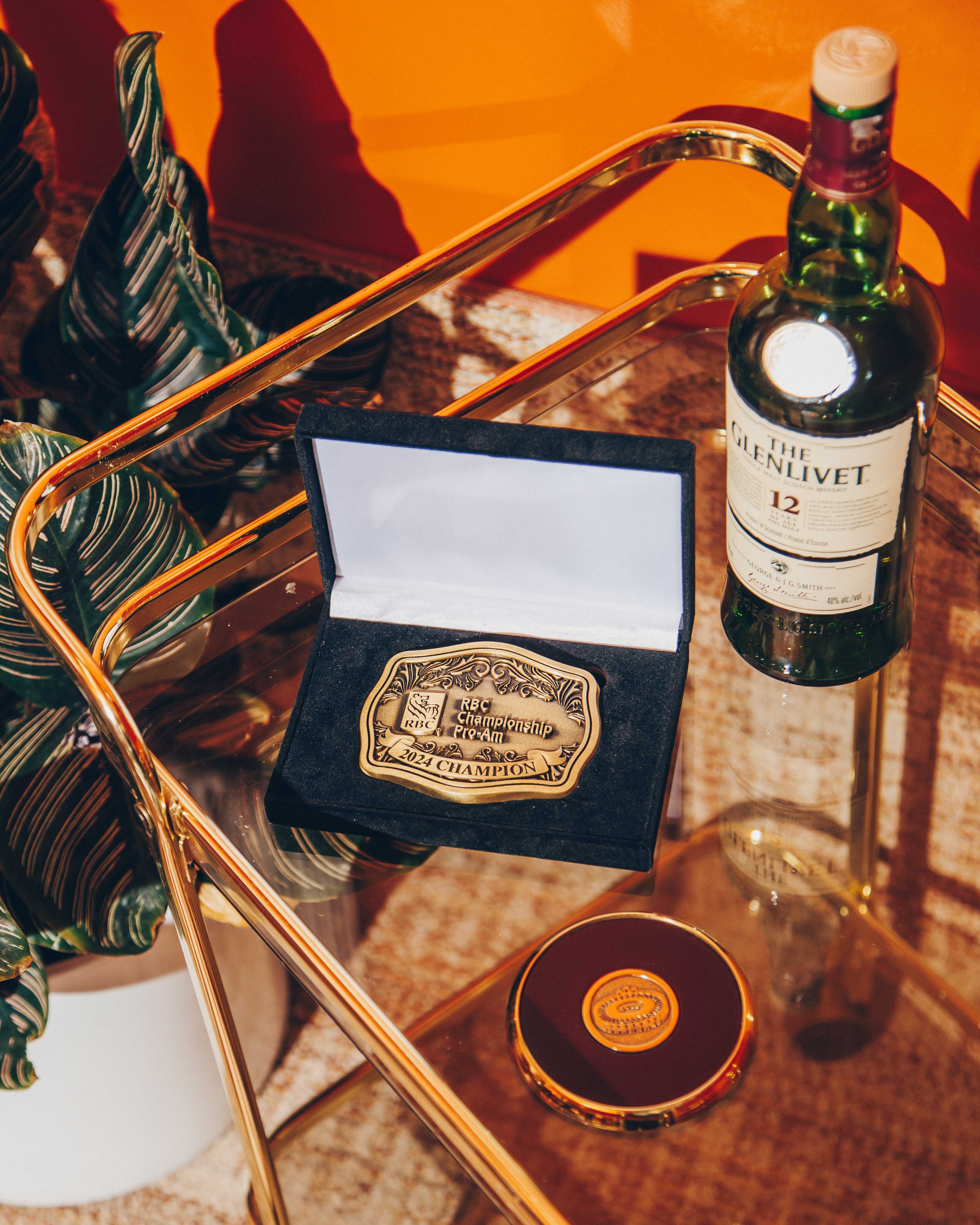 A glass and gold metal bar cart with a vibrant orange background. On the cart, there is a bottle of The Glenlivet 12-year-old Scotch whisky, a black velvet box containing a gold championship belt buckle, and a round, gold-rimmed object with a logo on top. The belt buckle is engraved with 'RBC Championship Pro-Am 2024 Champion.' The lighting is warm and casts distinct shadows, creating a dramatic and luxurious atmosphere. A potted plant with large, dark green leaves is partially visible on the left, adding a touch of nature to the scene.