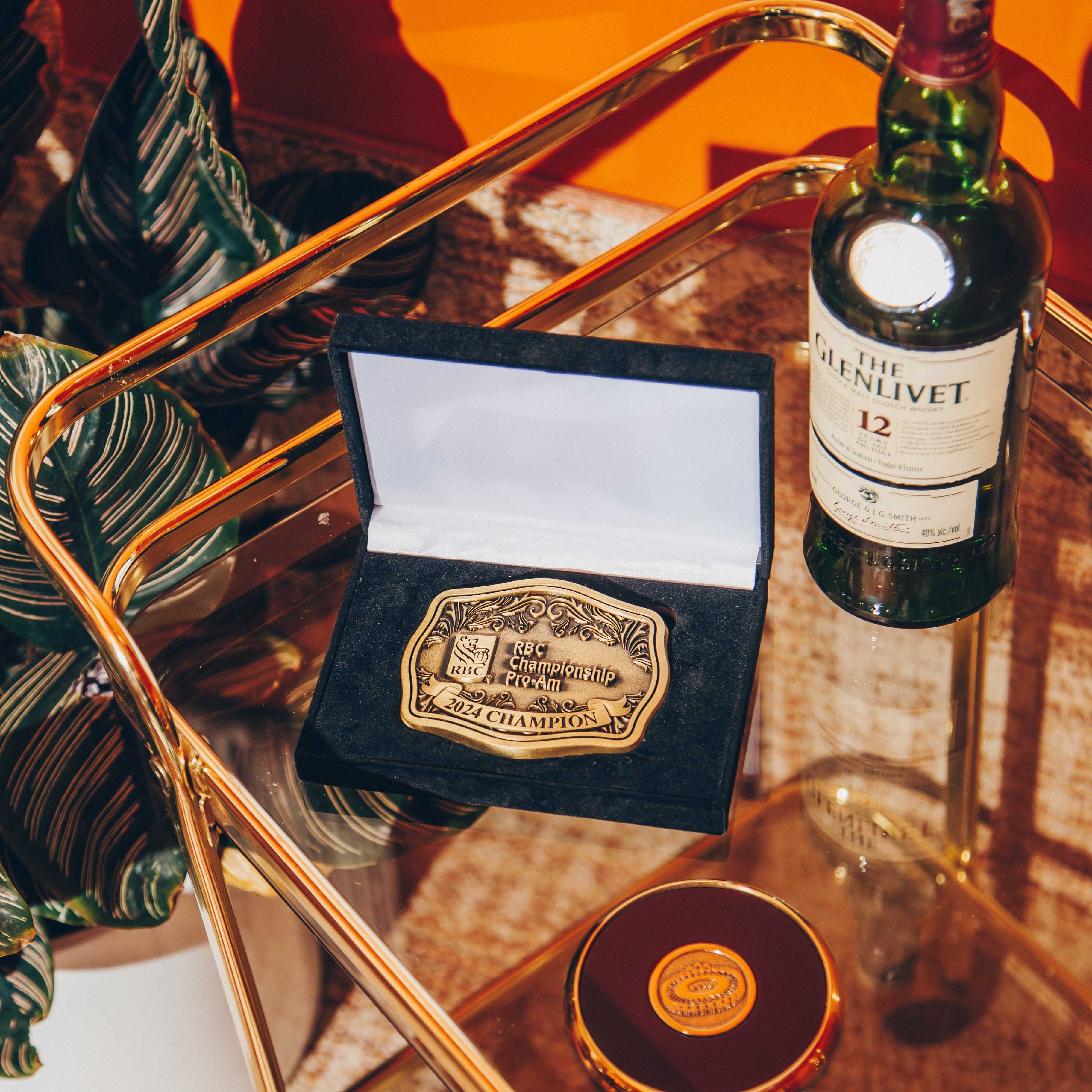 A glass and gold metal bar cart with a vibrant orange background. On the cart, there is a bottle of The Glenlivet 12-year-old Scotch whisky, a black velvet box containing a gold championship belt buckle, and a round, gold-rimmed object with a logo on top. The belt buckle is engraved with 'RBC Championship Pro-Am 2024 Champion.' The lighting is warm and casts distinct shadows, creating a dramatic and luxurious atmosphere. A potted plant with large, dark green leaves is partially visible on the left, adding a touch of nature to the scene.