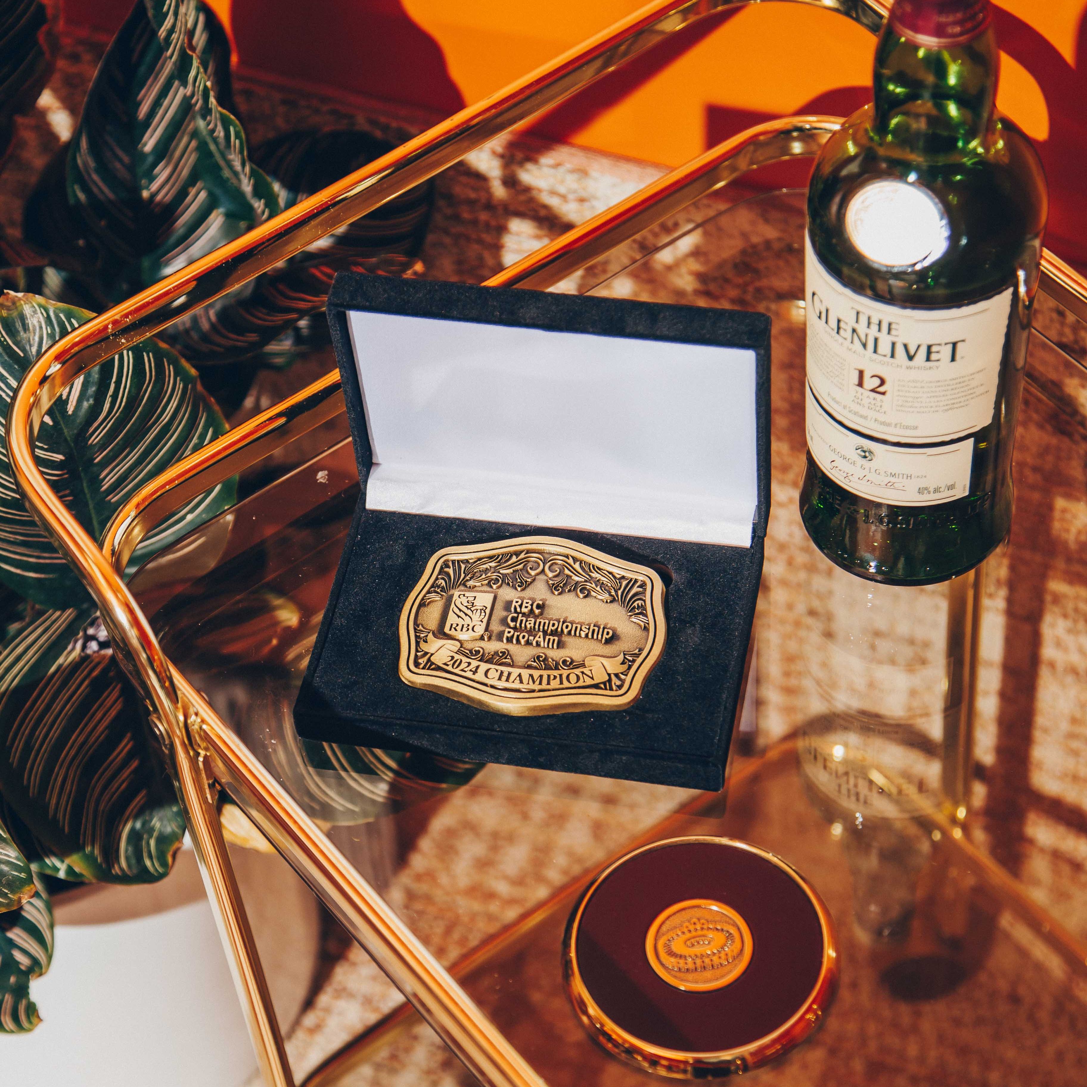A glass and gold metal bar cart with a vibrant orange background. On the cart, there is a bottle of The Glenlivet 12-year-old Scotch whisky, a black velvet box containing a gold championship belt buckle, and a round, gold-rimmed object with a logo on top. The belt buckle is engraved with 'RBC Championship Pro-Am 2024 Champion.' The lighting is warm and casts distinct shadows, creating a dramatic and luxurious atmosphere. A potted plant with large, dark green leaves is partially visible on the left, adding a touch of nature to the scene.