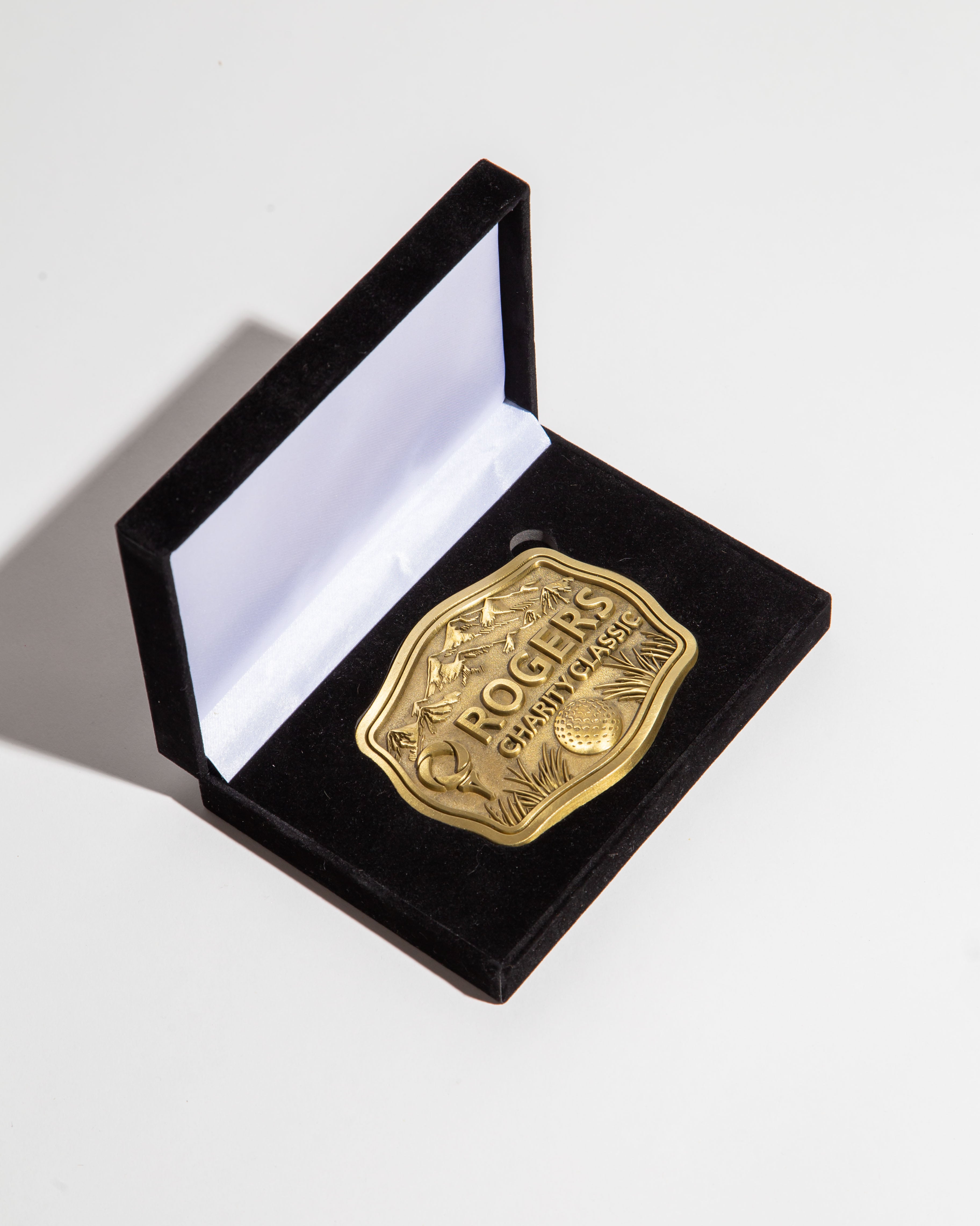 A gold-colored belt-buckle placed inside a black velvet presentation box. The buckle is intricately designed with the text ROGERS CHARITY CLASSIC prominently displayed above imagery of a golf ball and landscape.