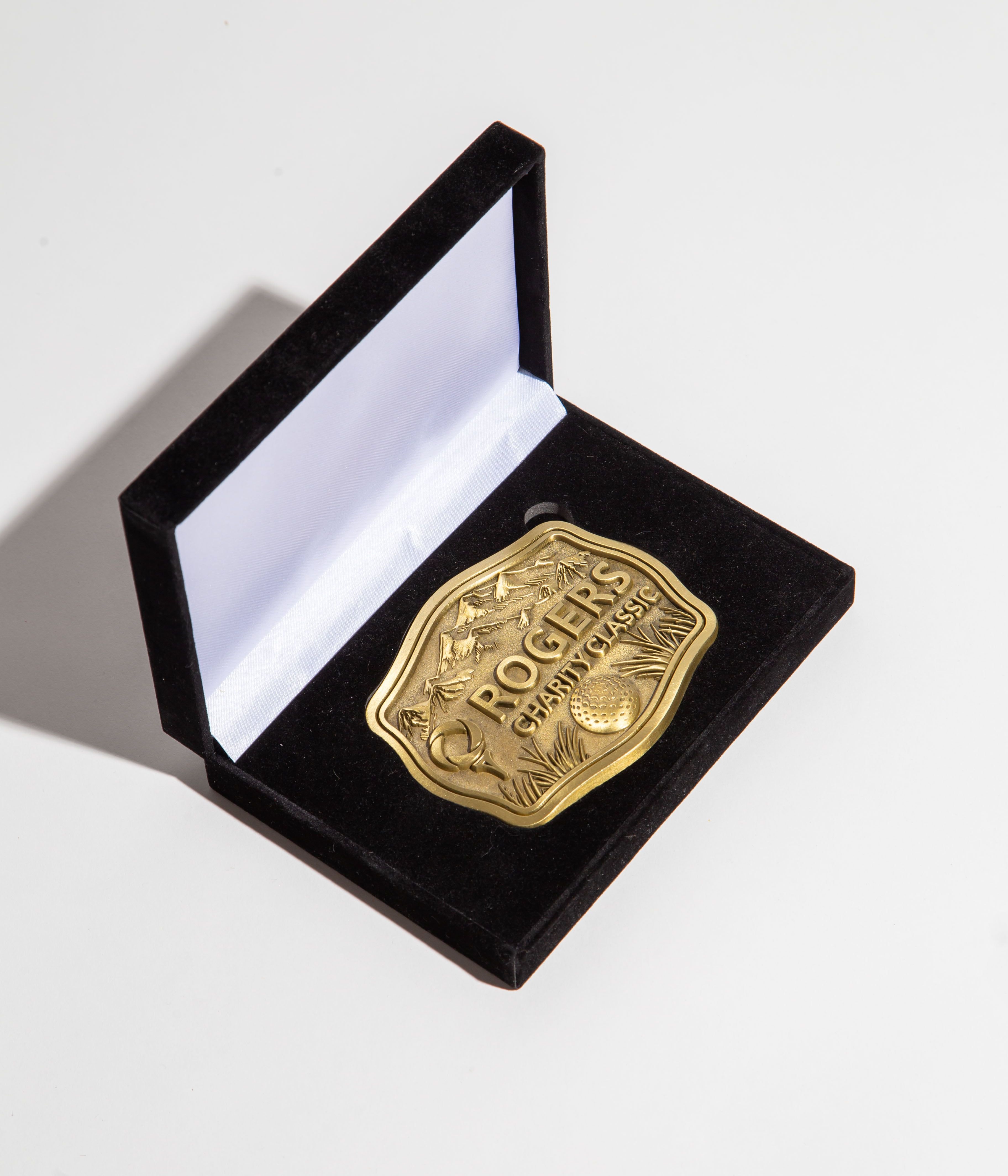 A gold-colored belt-buckle placed inside a black velvet presentation box. The buckle is intricately designed with the text ROGERS CHARITY CLASSIC prominently displayed above imagery of a golf ball and landscape.