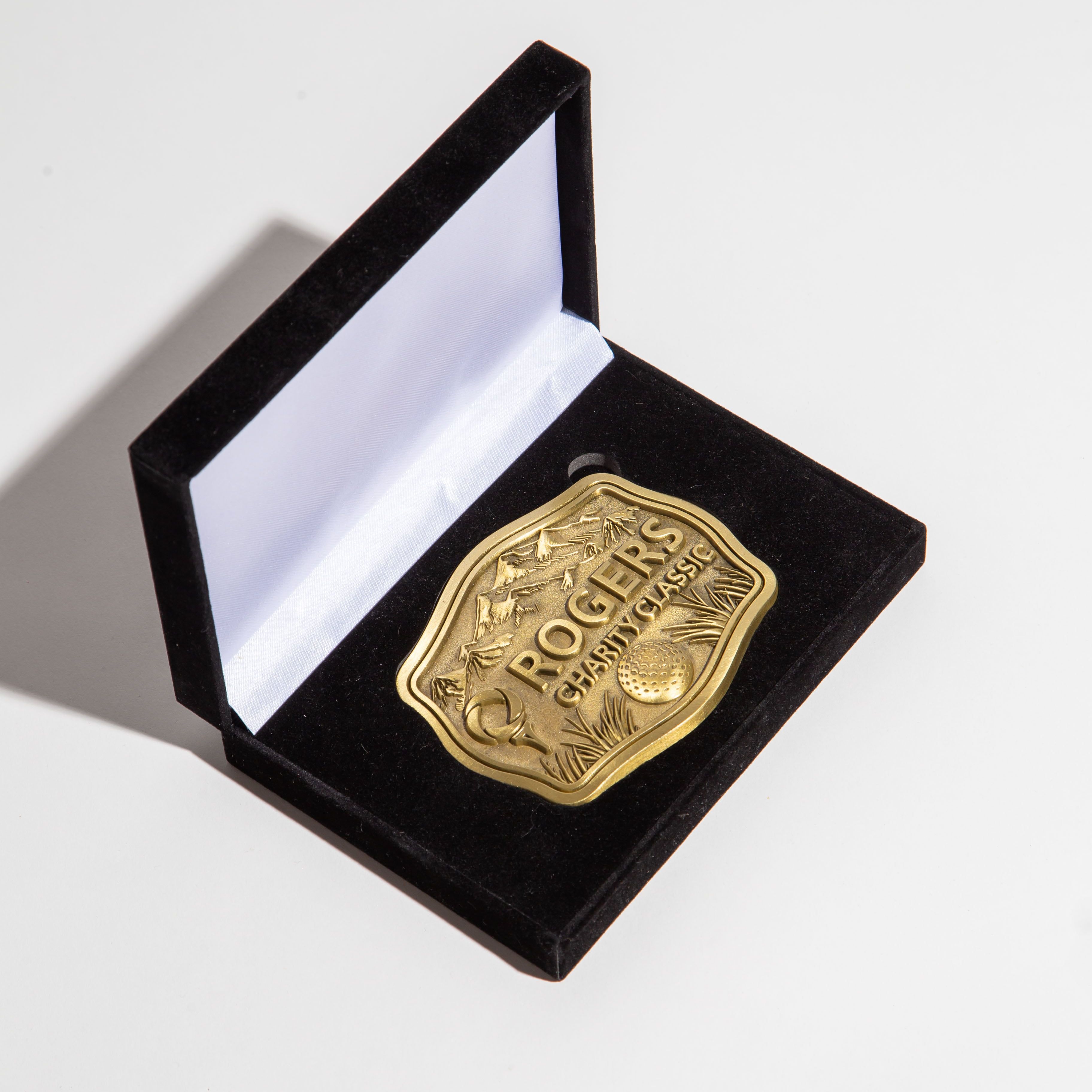 A gold-colored belt-buckle placed inside a black velvet presentation box. The buckle is intricately designed with the text ROGERS CHARITY CLASSIC prominently displayed above imagery of a golf ball and landscape.