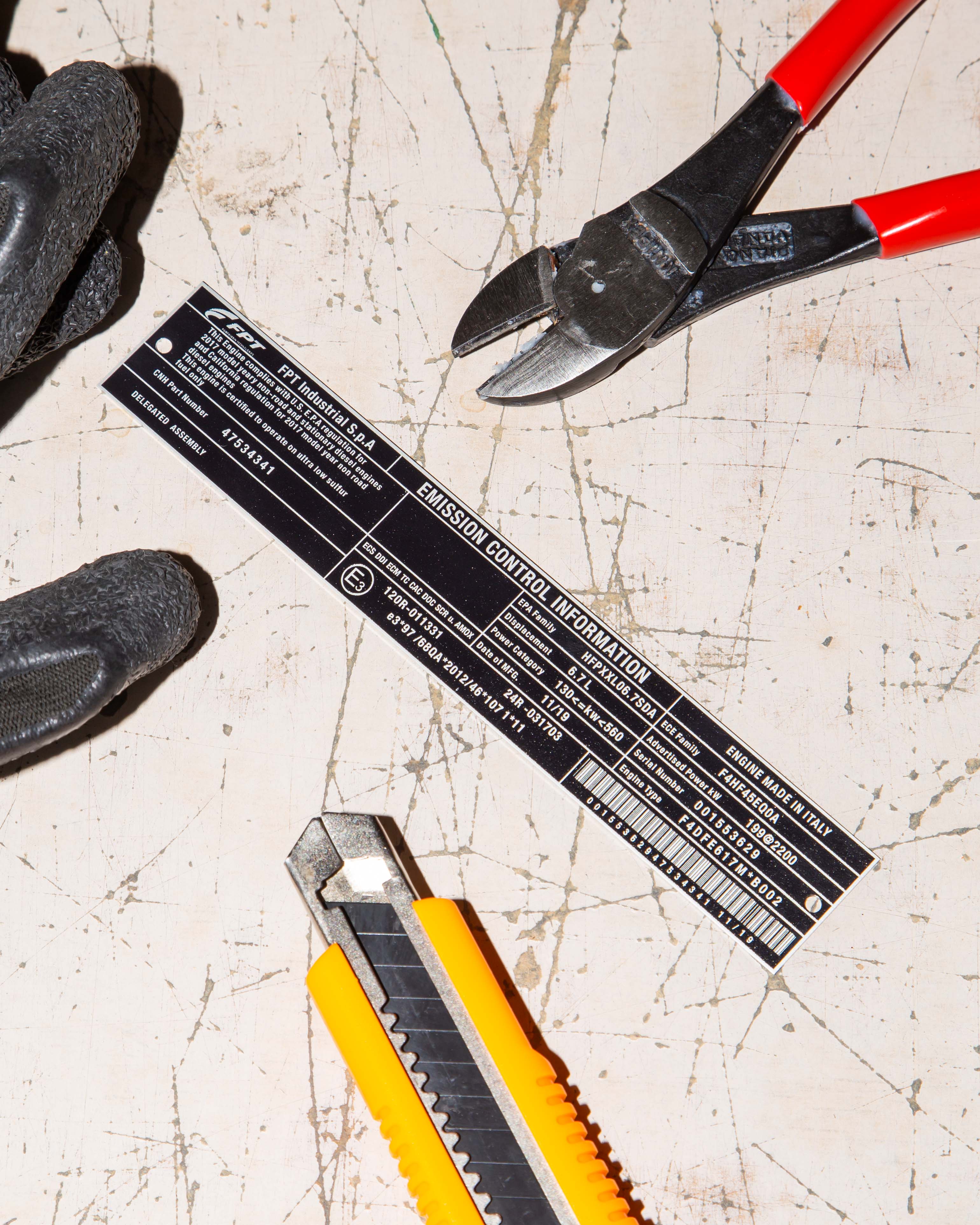 Close-up of a utility knife, pliers, and emission control label on a scratched surface.