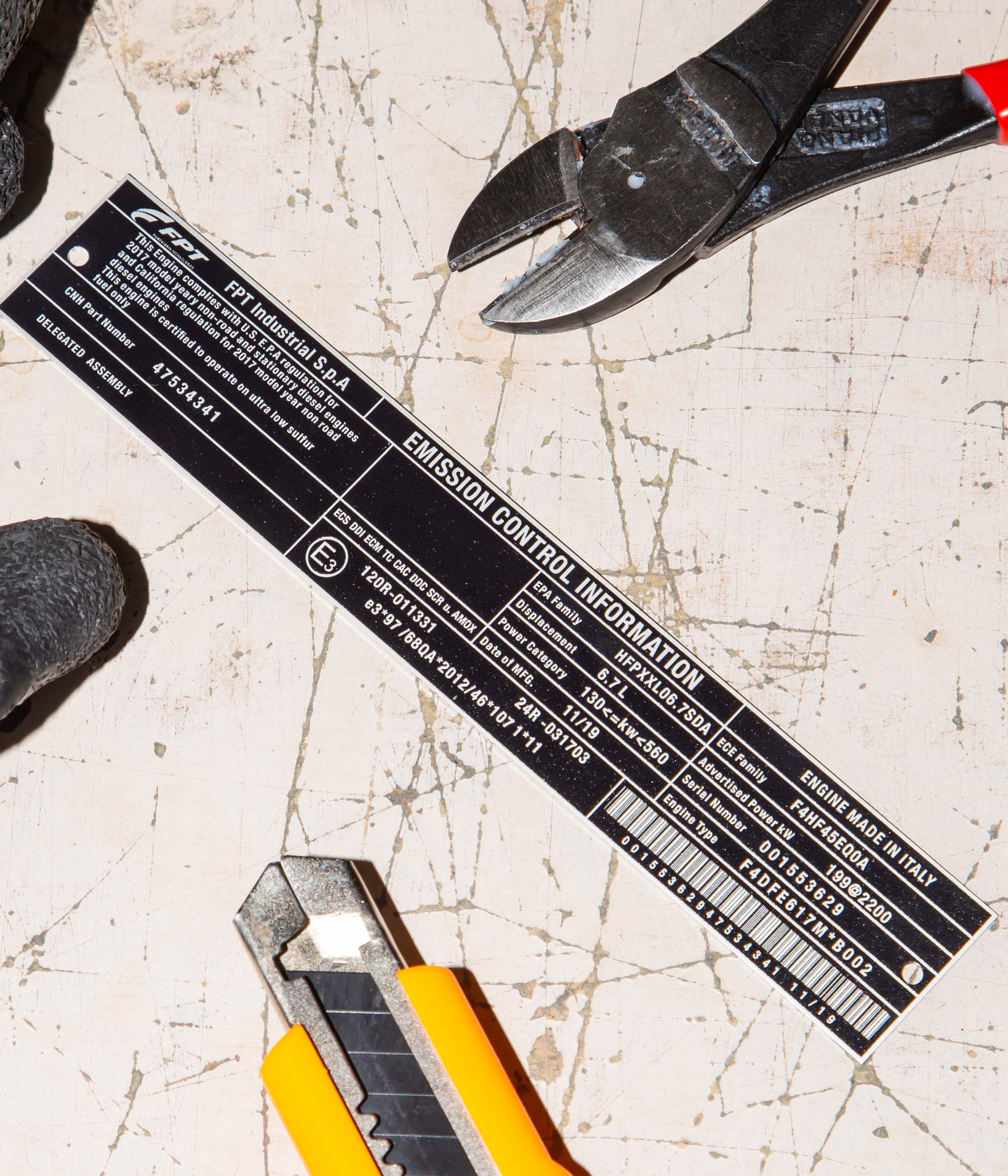 Close-up of a utility knife, pliers, and emission control label on a scratched surface.