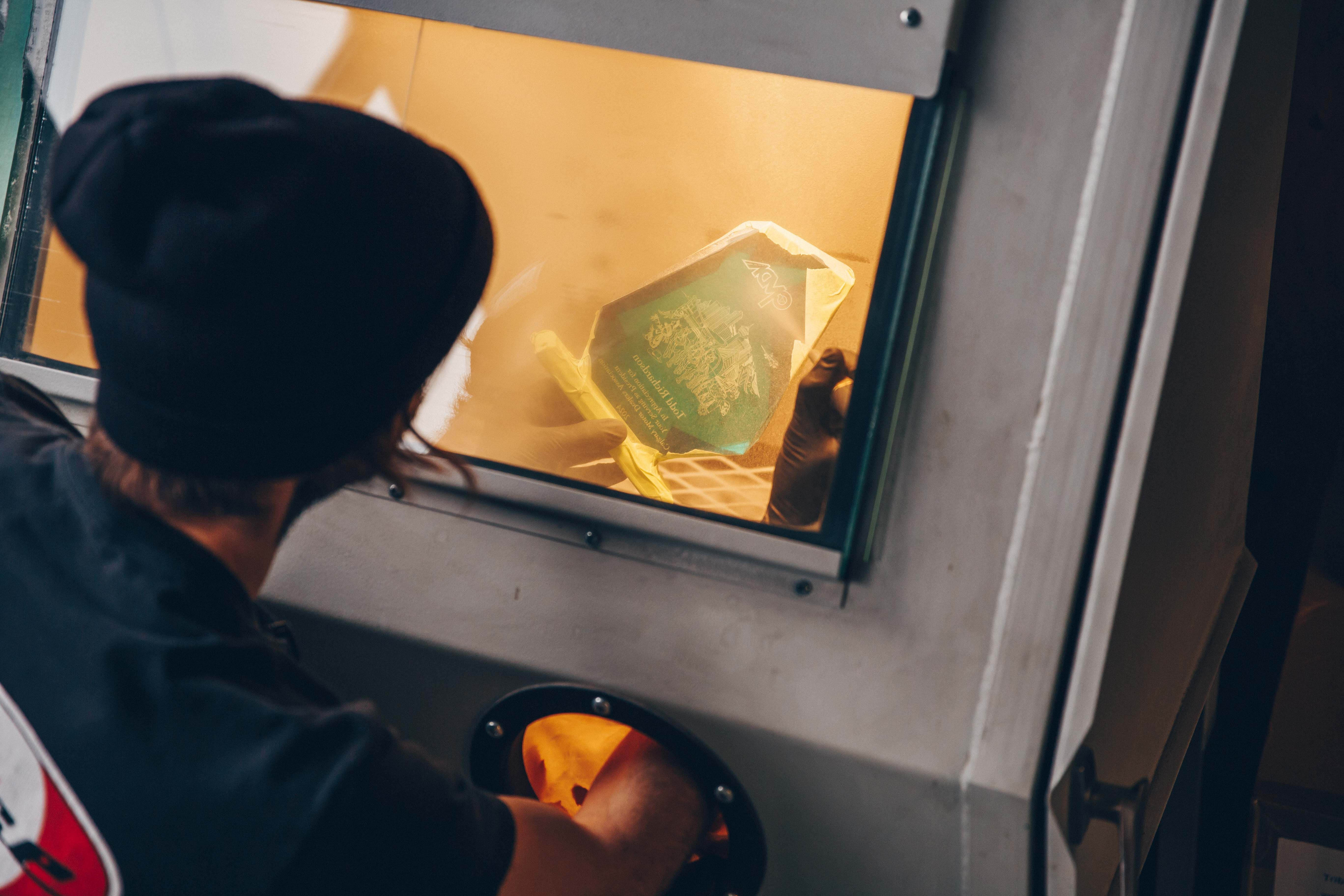 Close-up view of a person working inside a sandblasting cabinet, holding a glass piece with taped sections for precise engraving.