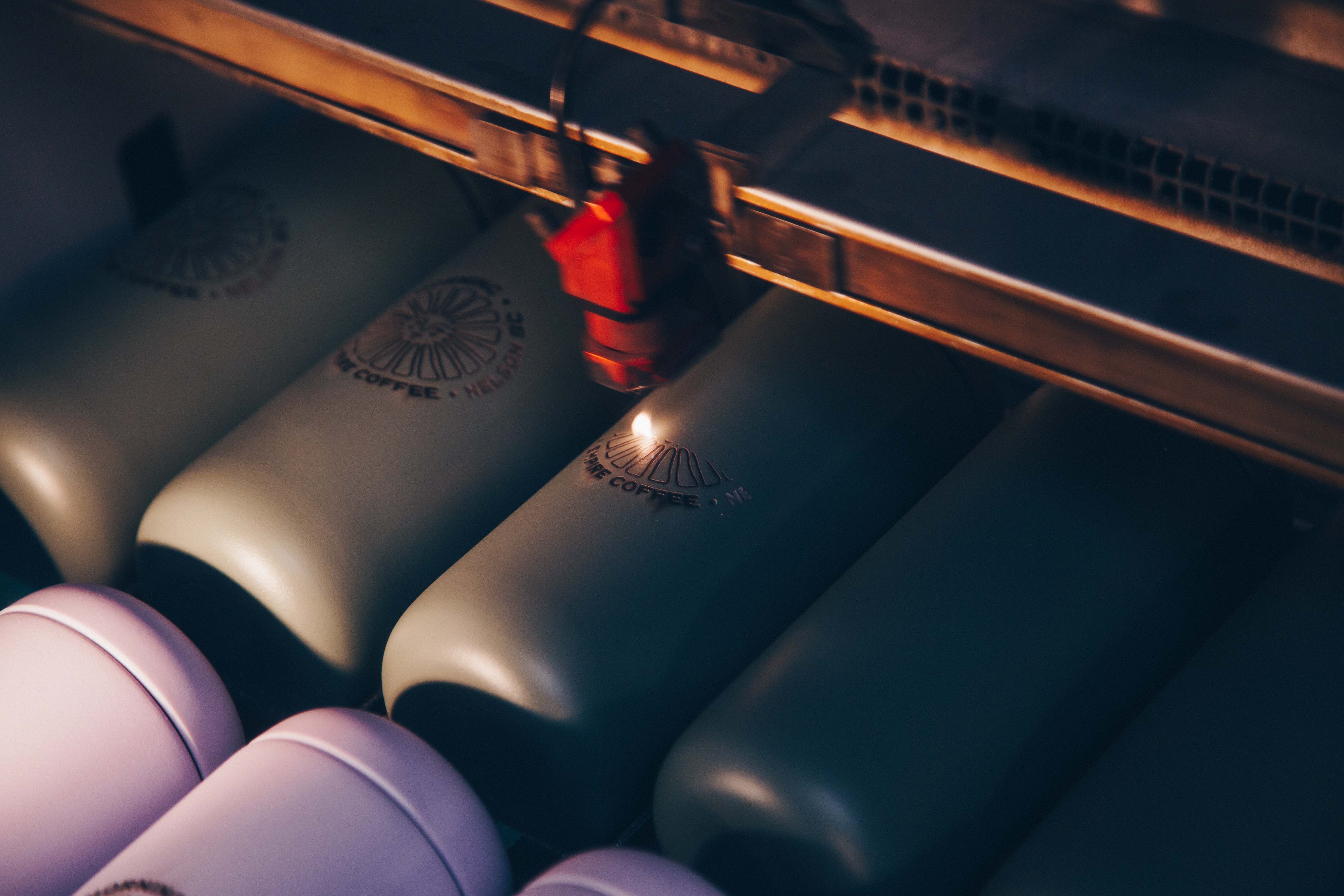 A laser engraving machine etching the "Good Morning Coffee, Nelson BC" logo onto green drinkware. The machine’s precision highlights the quality of custom branding for coffee tumblers and promotional products.