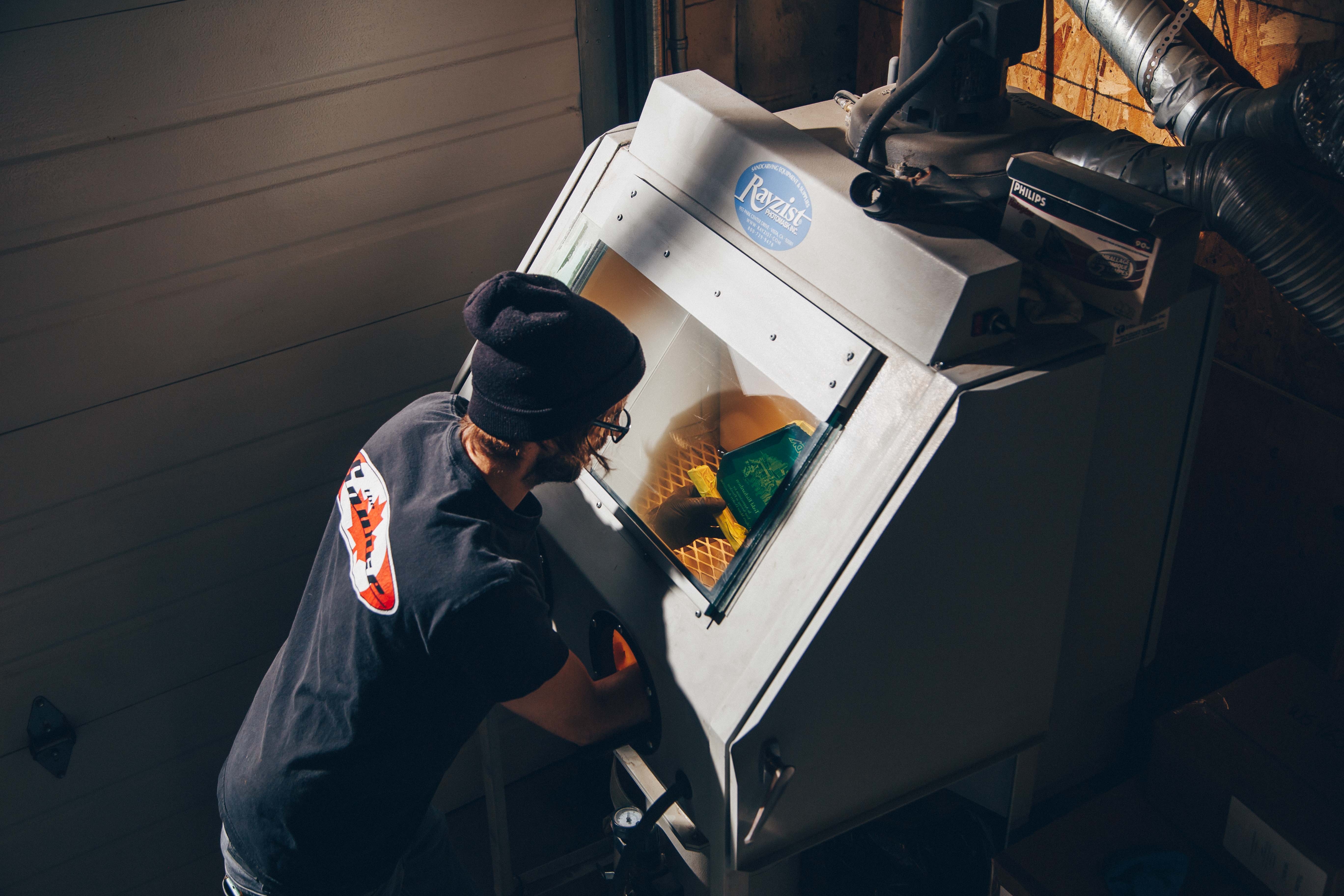 A person working at a sandblasting cabinet, carefully engraving designs onto a glass award inside a lit workspace.