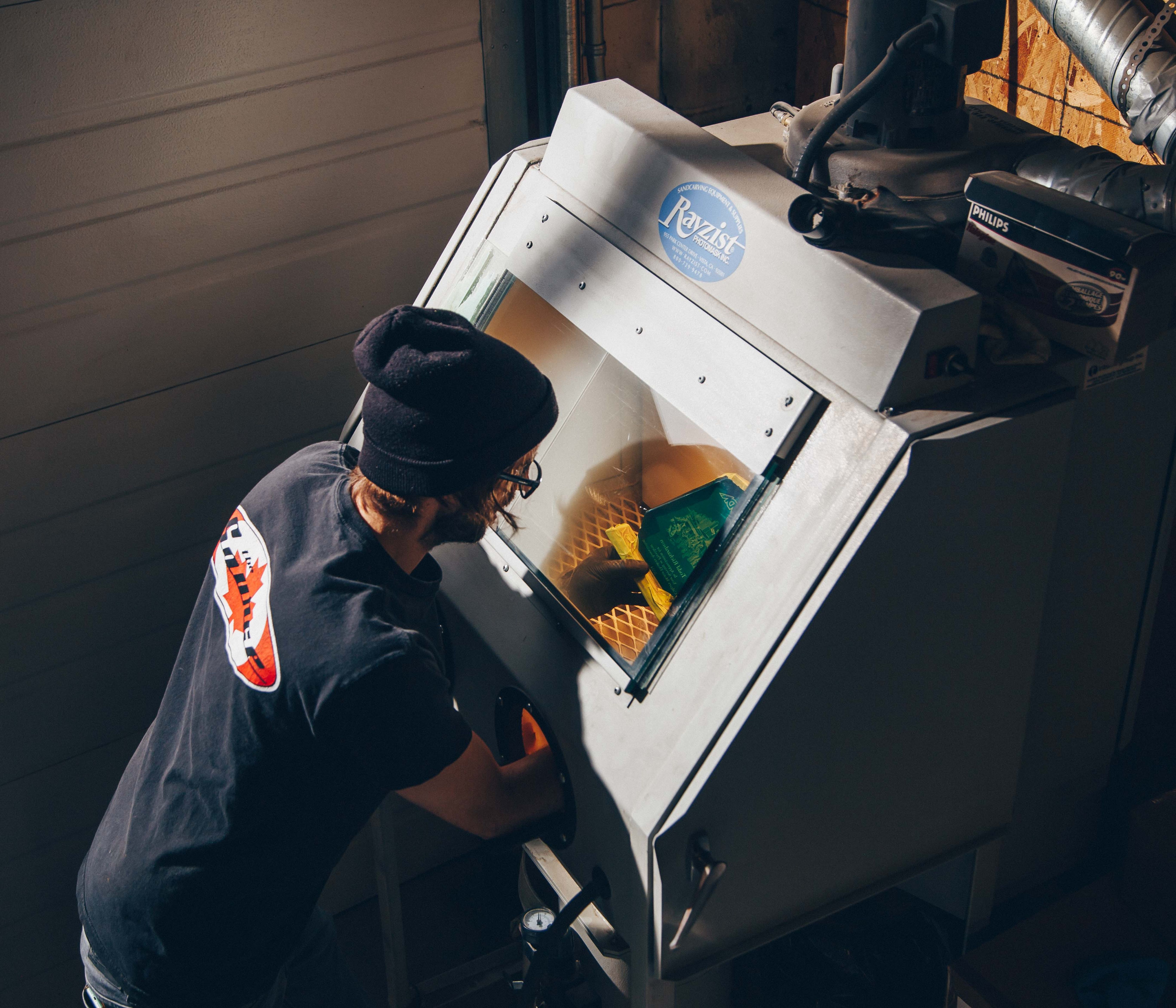 A person working at a sandblasting cabinet, carefully engraving designs onto a glass award inside a lit workspace.