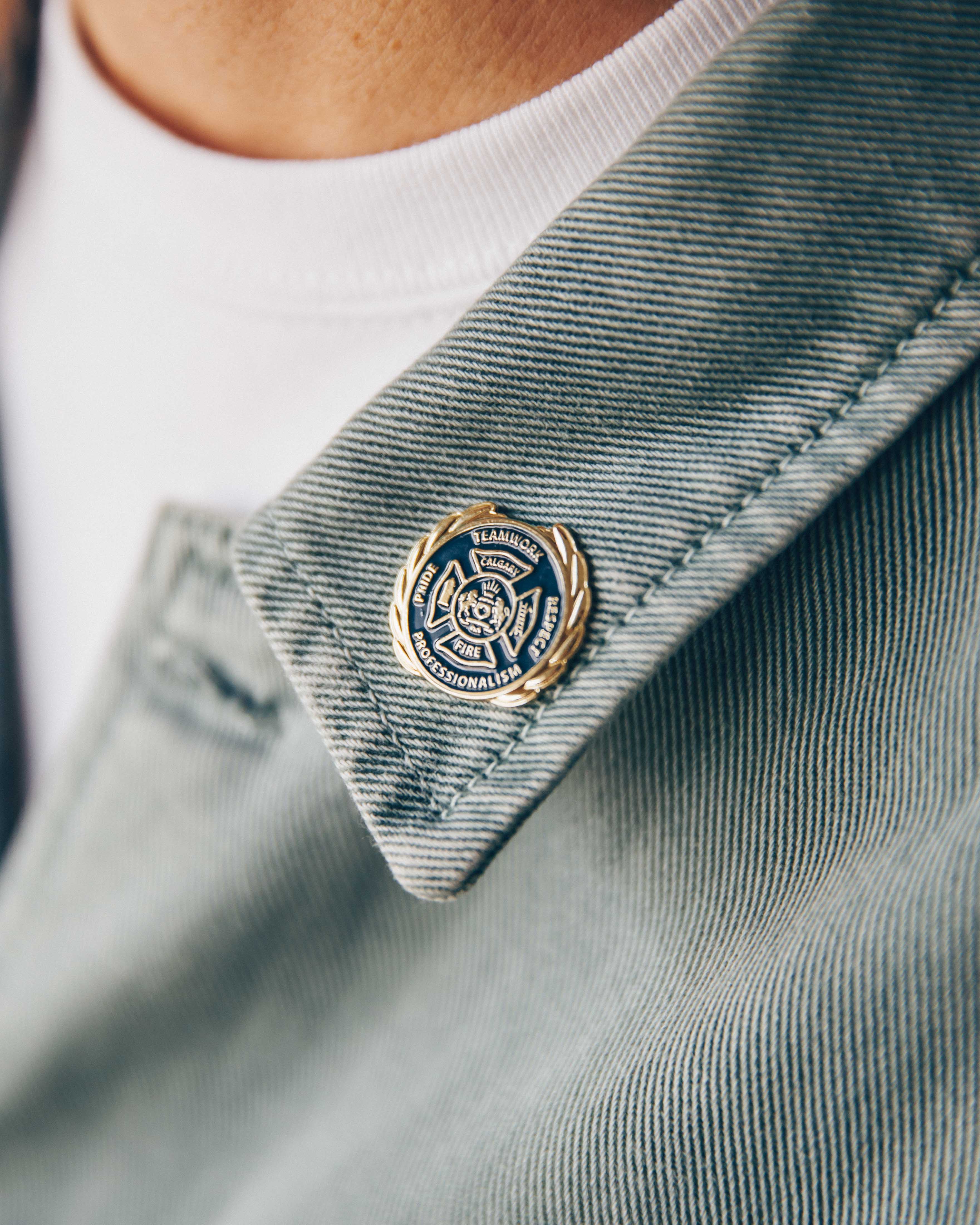 Close-up of a circular enamel pin on grey denim jacket collar, with white t-shirt visible underneath.