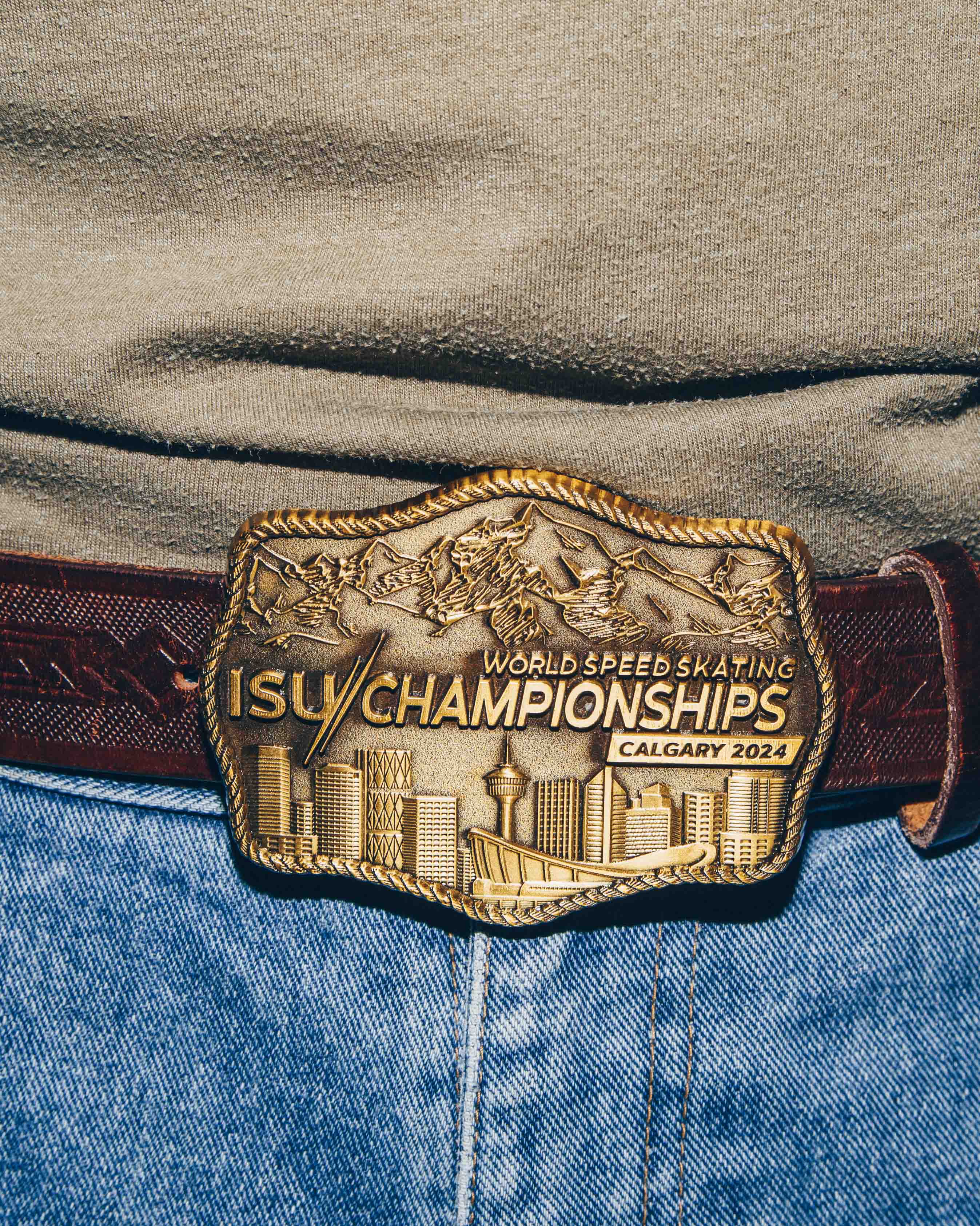 A close-up view of a belt buckle worn on a pair of blue jeans. The buckle is intricately designed with a gold finish, depicting a city skyline and mountain range. It is engraved with the text ISU World Speed Skating Championships Calgary 2024. The belt is made of dark brown leather with a textured pattern.