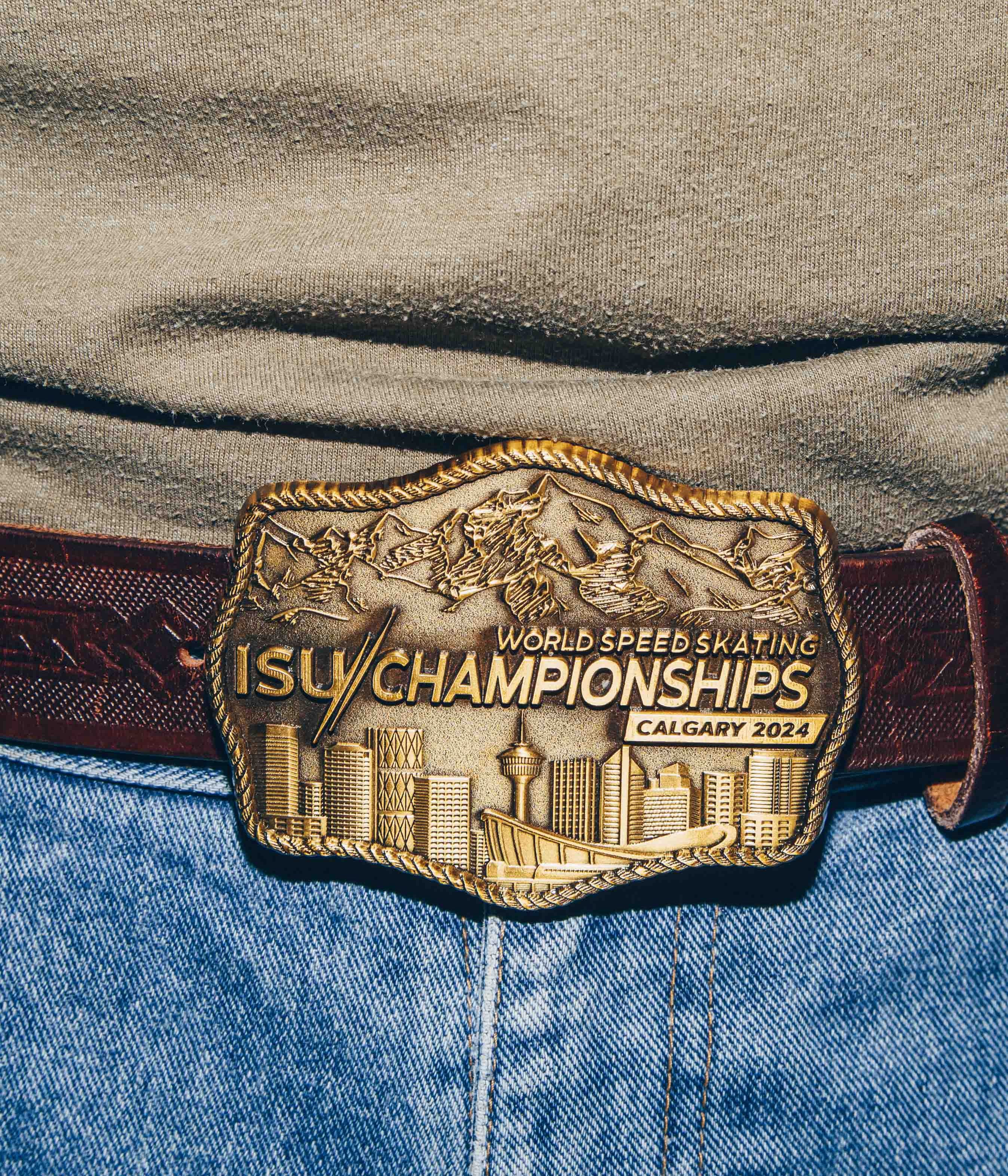 A close-up view of a belt buckle worn on a pair of blue jeans. The buckle is intricately designed with a gold finish, depicting a city skyline and mountain range. It is engraved with the text ISU World Speed Skating Championships Calgary 2024. The belt is made of dark brown leather with a textured pattern.