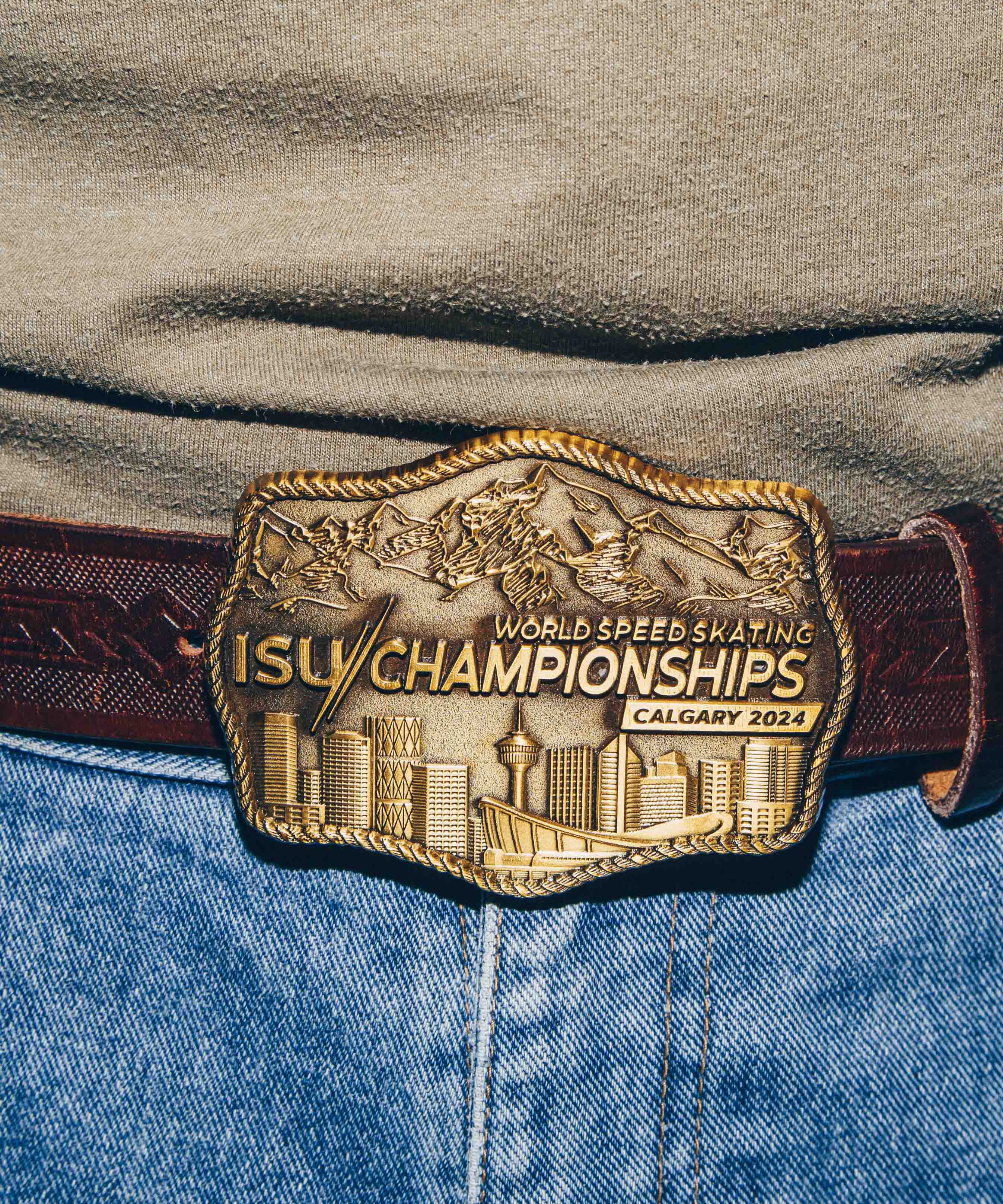 A close-up view of a belt buckle worn on a pair of blue jeans. The buckle is intricately designed with a gold finish, depicting a city skyline and mountain range. It is engraved with the text ISU World Speed Skating Championships Calgary 2024. The belt is made of dark brown leather with a textured pattern.
