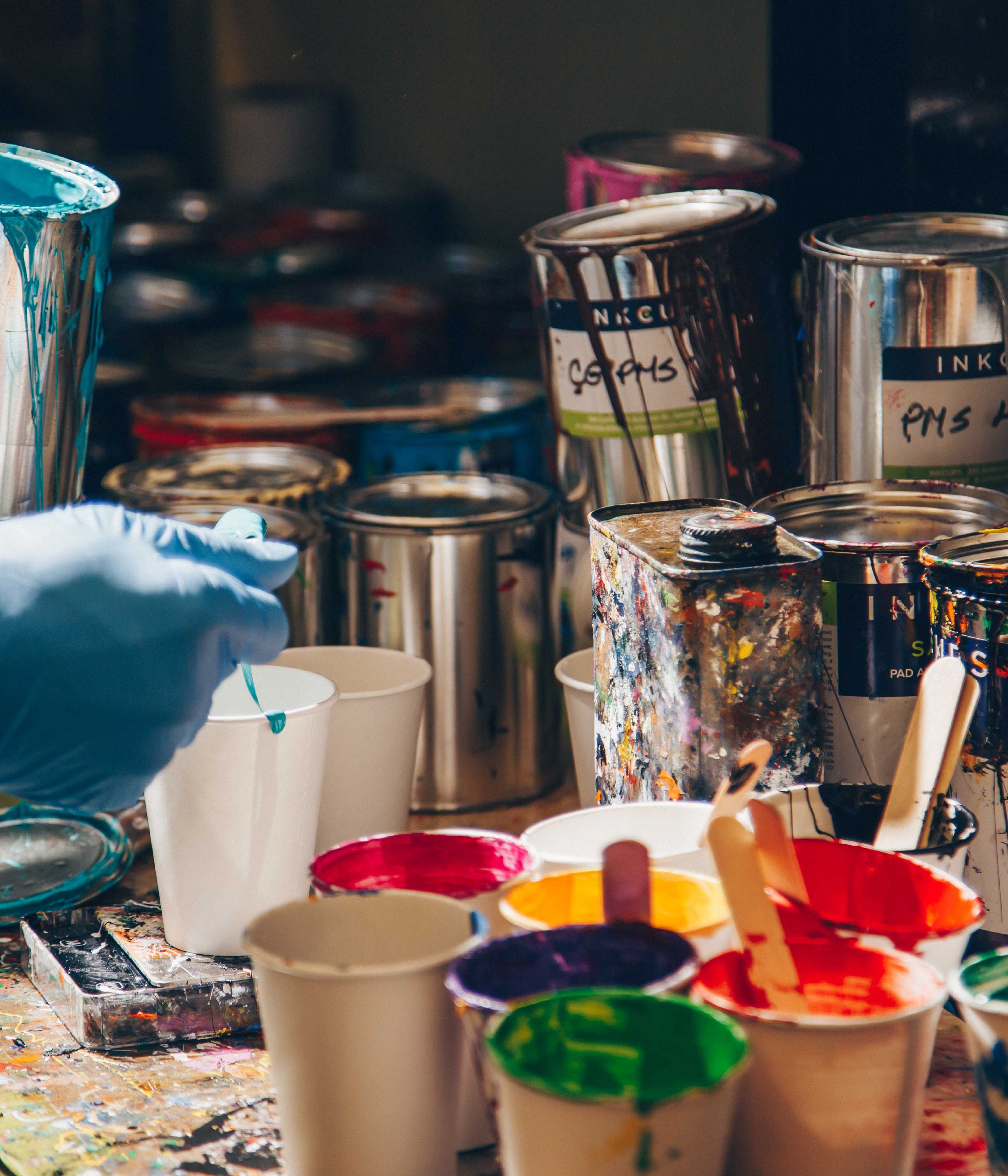 A paint preparation area in a pad printing shop featuring colorful ink cups, metal paint cans, and mixing tools. A worker wearing blue gloves stirs turquoise ink, preparing custom colors for pad printing designs on various products.