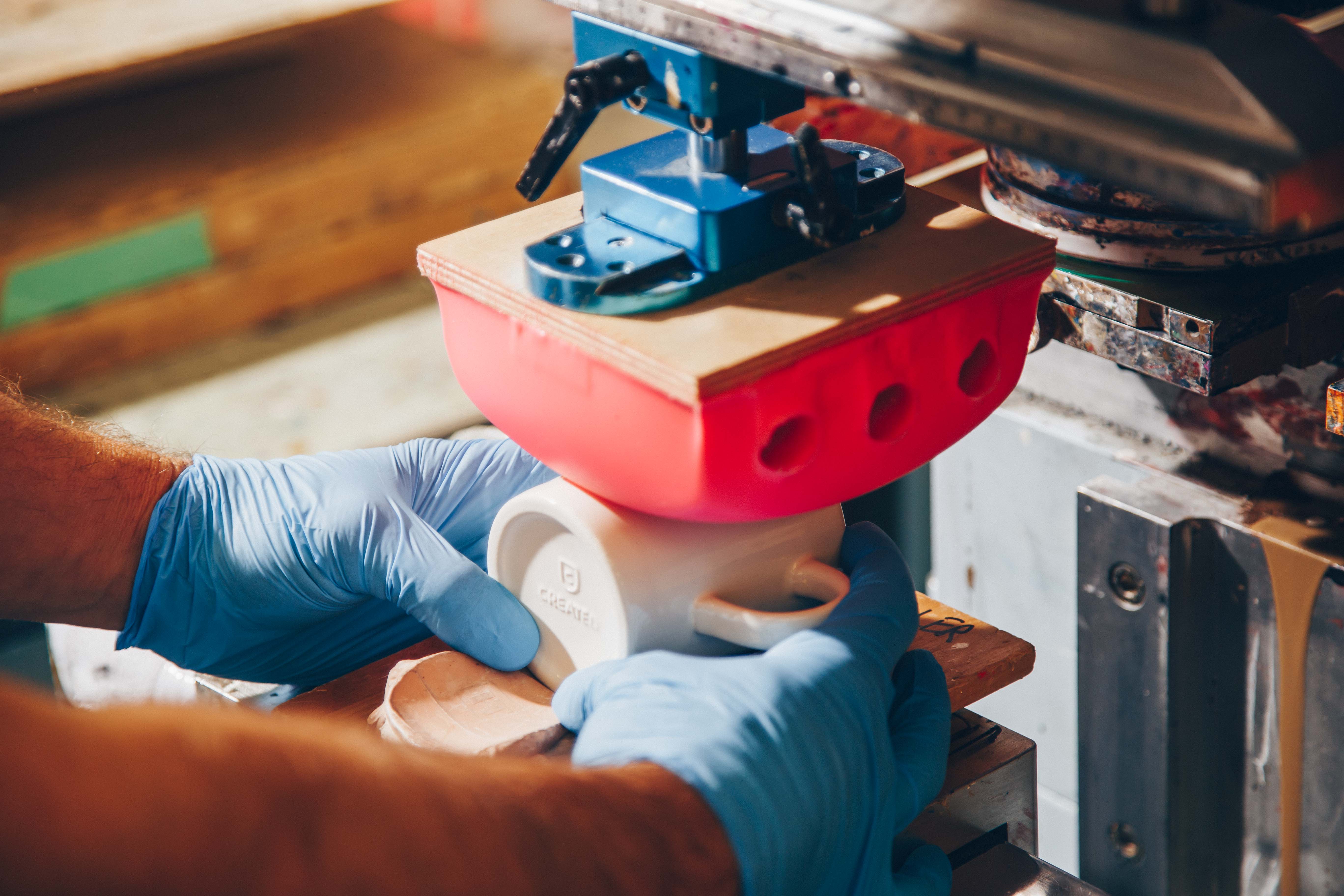 A close-up of a pad printing process where a red silicone pad transfers custom artwork onto a white ceramic mug. A worker wearing blue gloves carefully aligns the mug under the machine for precision branding.