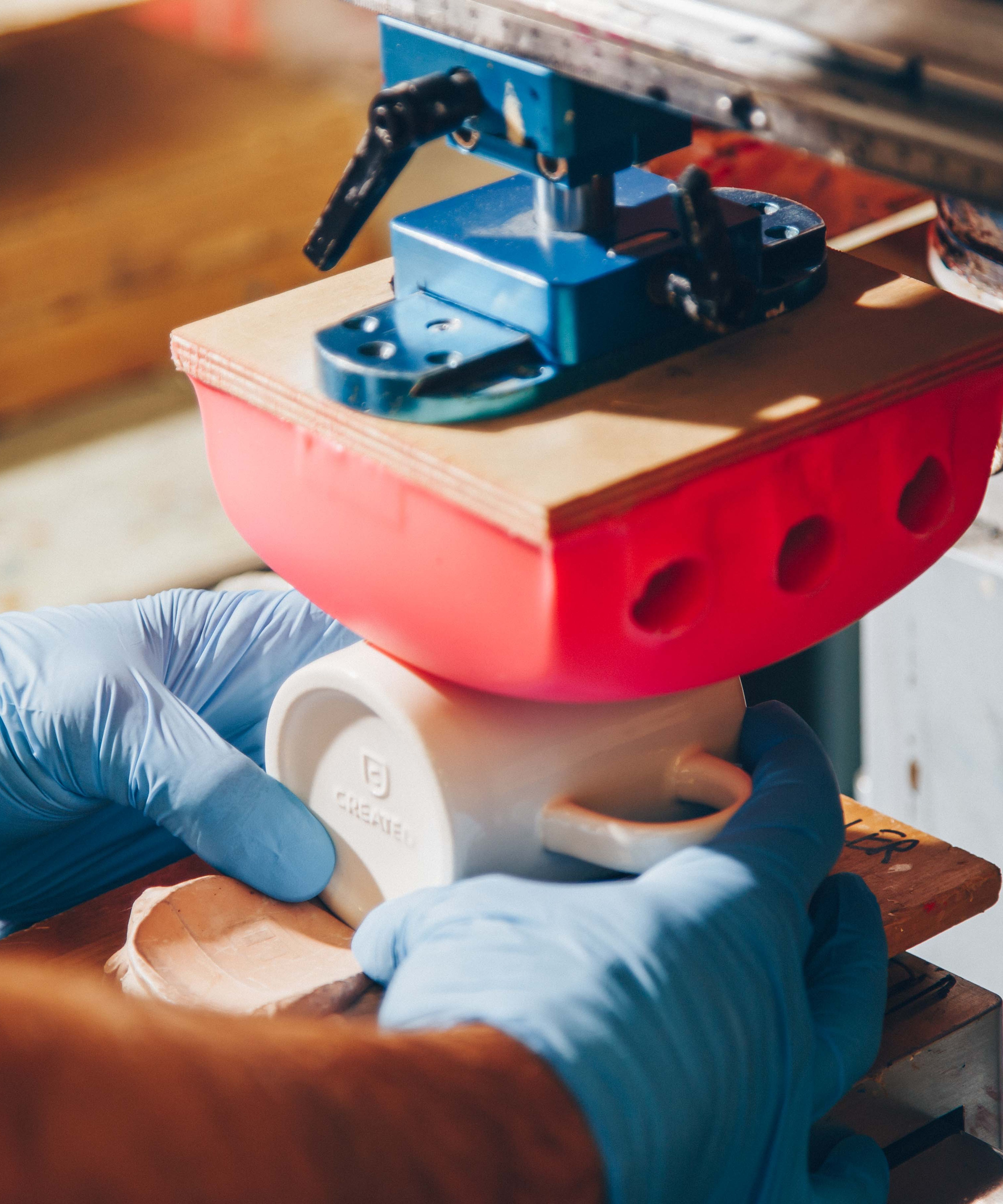A close-up of a pad printing process where a red silicone pad transfers custom artwork onto a white ceramic mug. A worker wearing blue gloves carefully aligns the mug under the machine for precision branding.