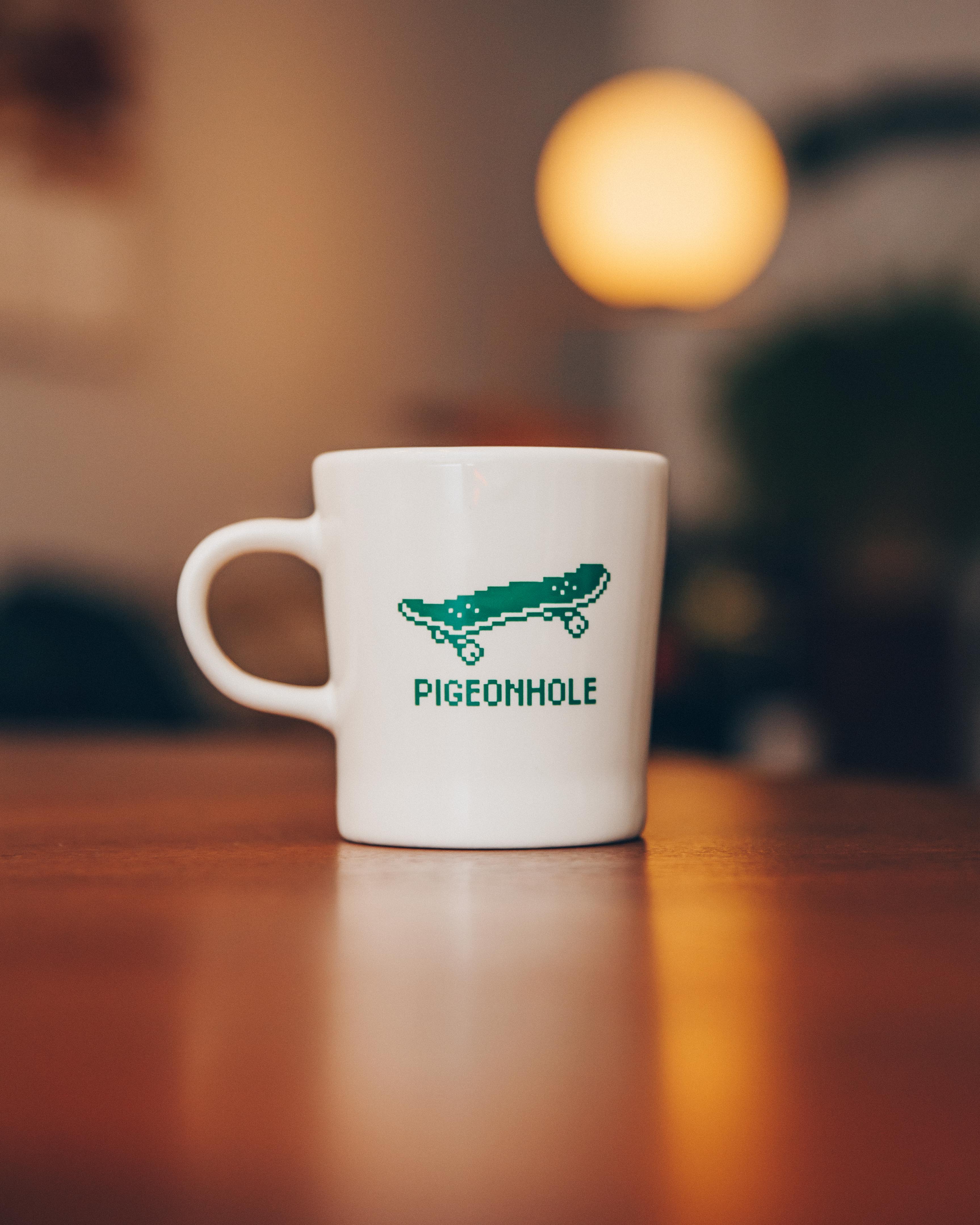 White mug with pixelated skateboard design and 'Pigeonhole' text on a wooden table.