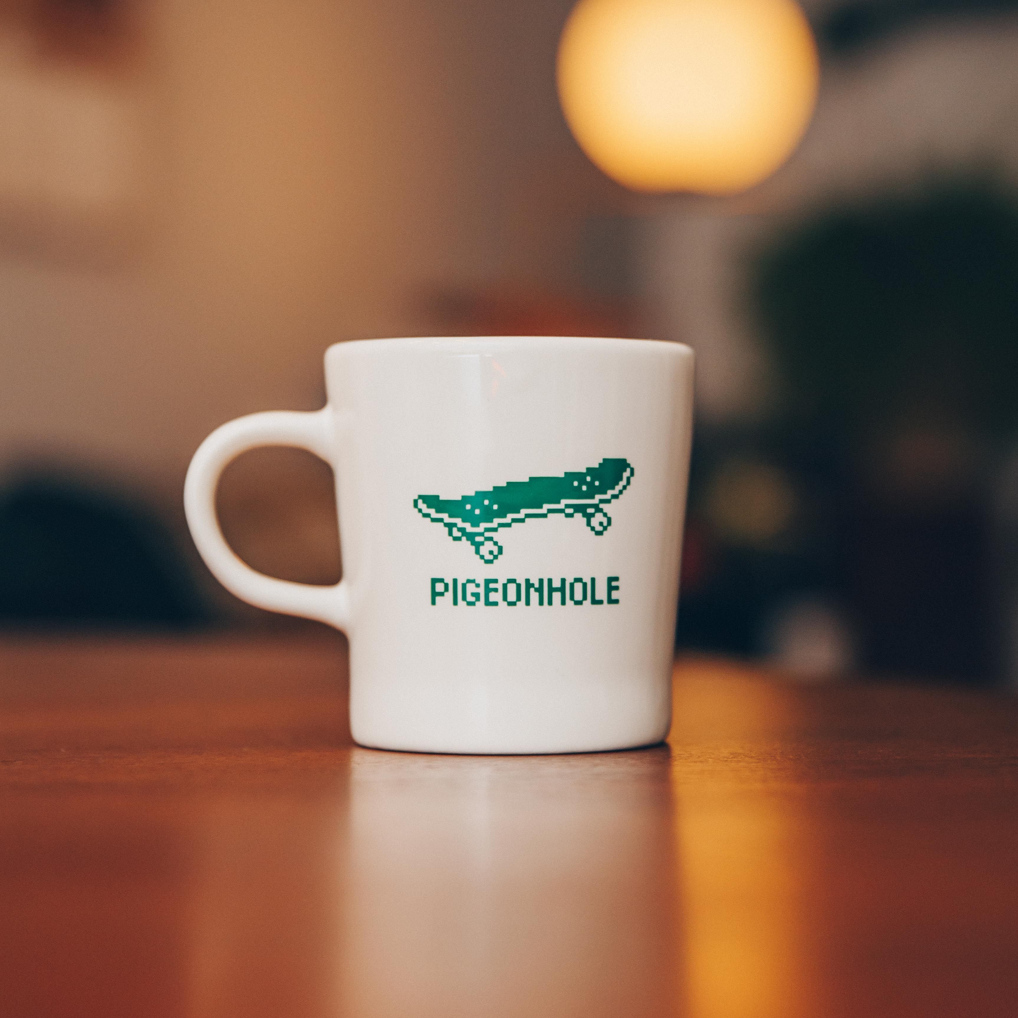 White mug with pixelated skateboard design and 'Pigeonhole' text on a wooden table.