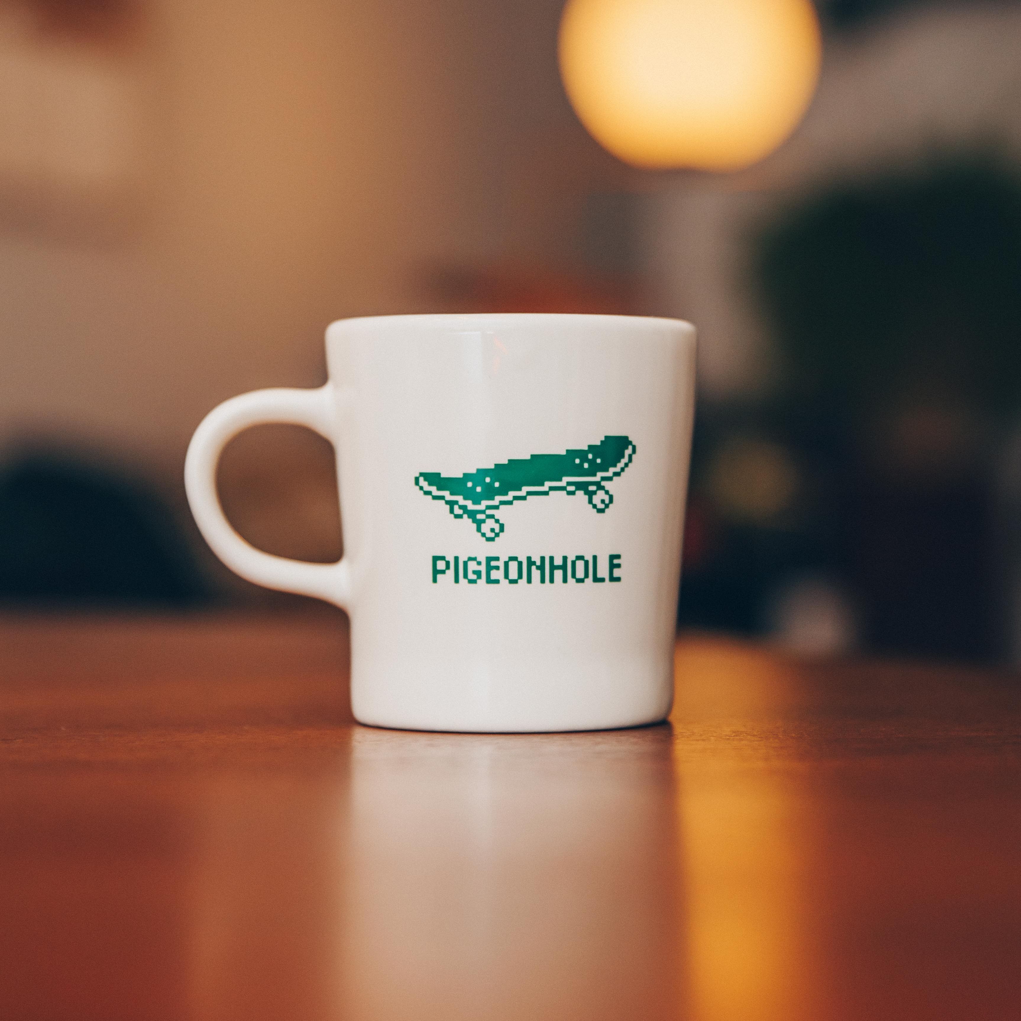 White mug with pixelated skateboard design and 'Pigeonhole' text on a wooden table.
