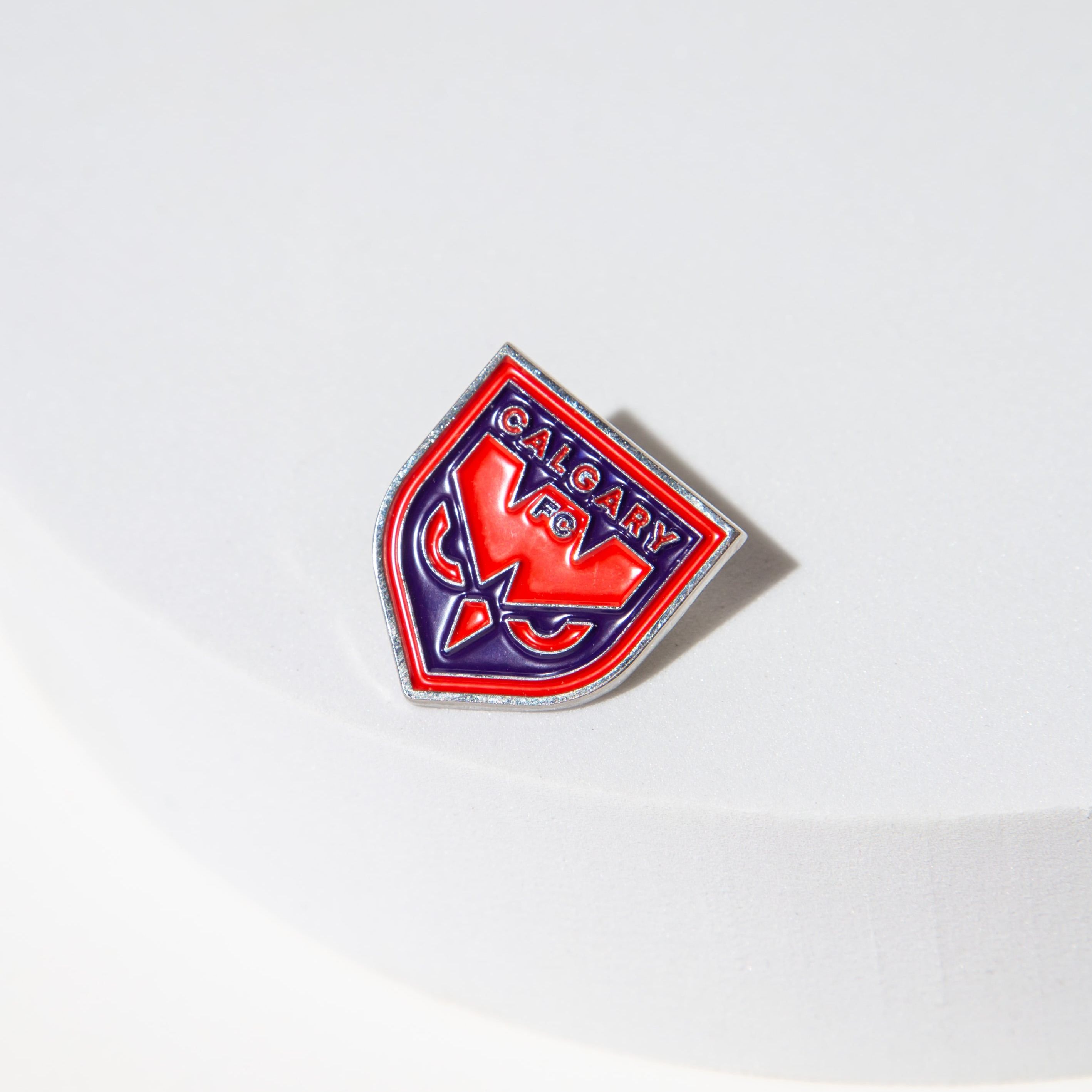 Red and blue Calgary FC enamel pin on a white surface.