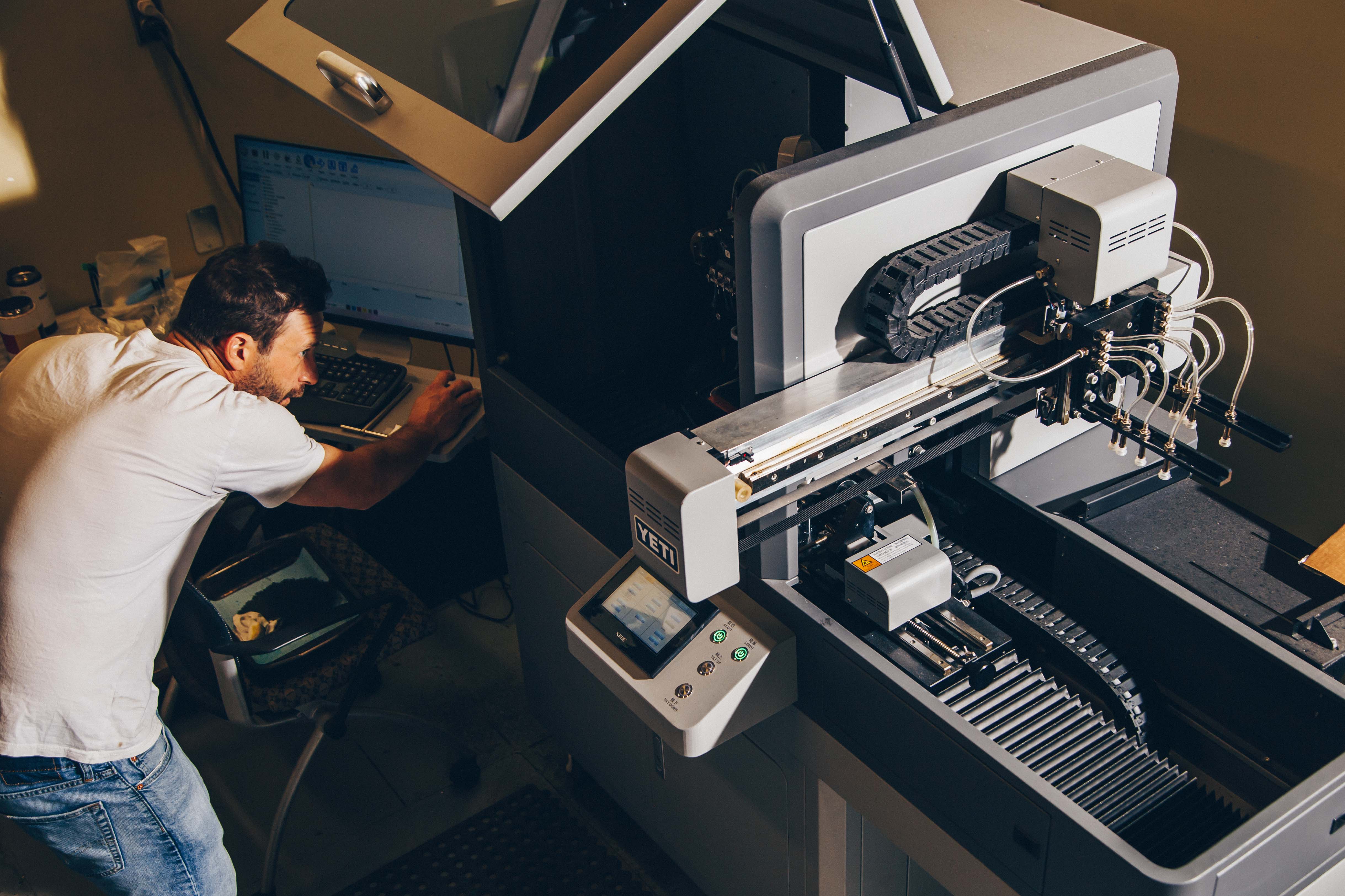 A person leans over a computer to manage a digital rotary printing machine, with components ready for customizing cylindrical drinkware.