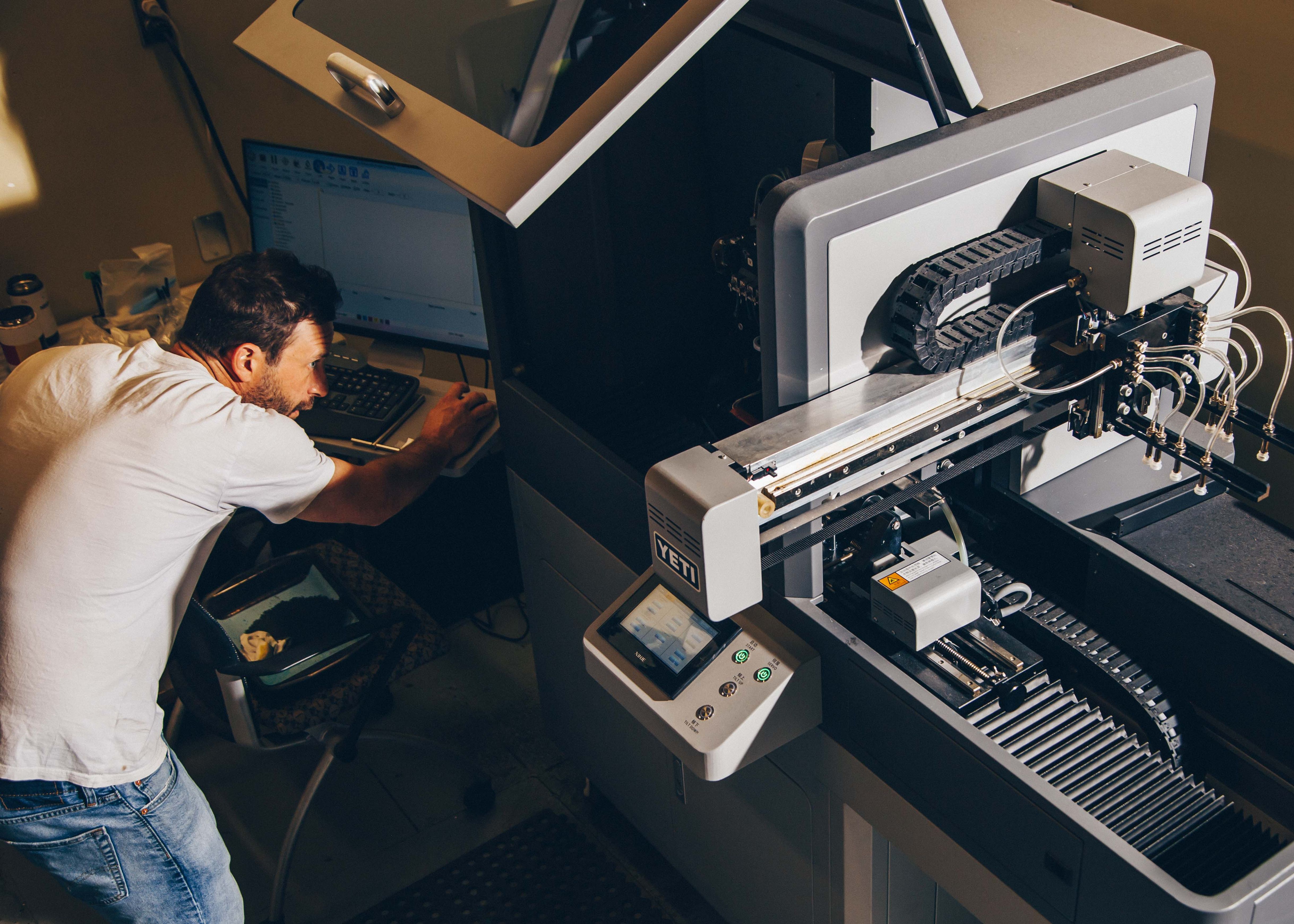 A person leans over a computer to manage a digital rotary printing machine, with components ready for customizing cylindrical drinkware.
