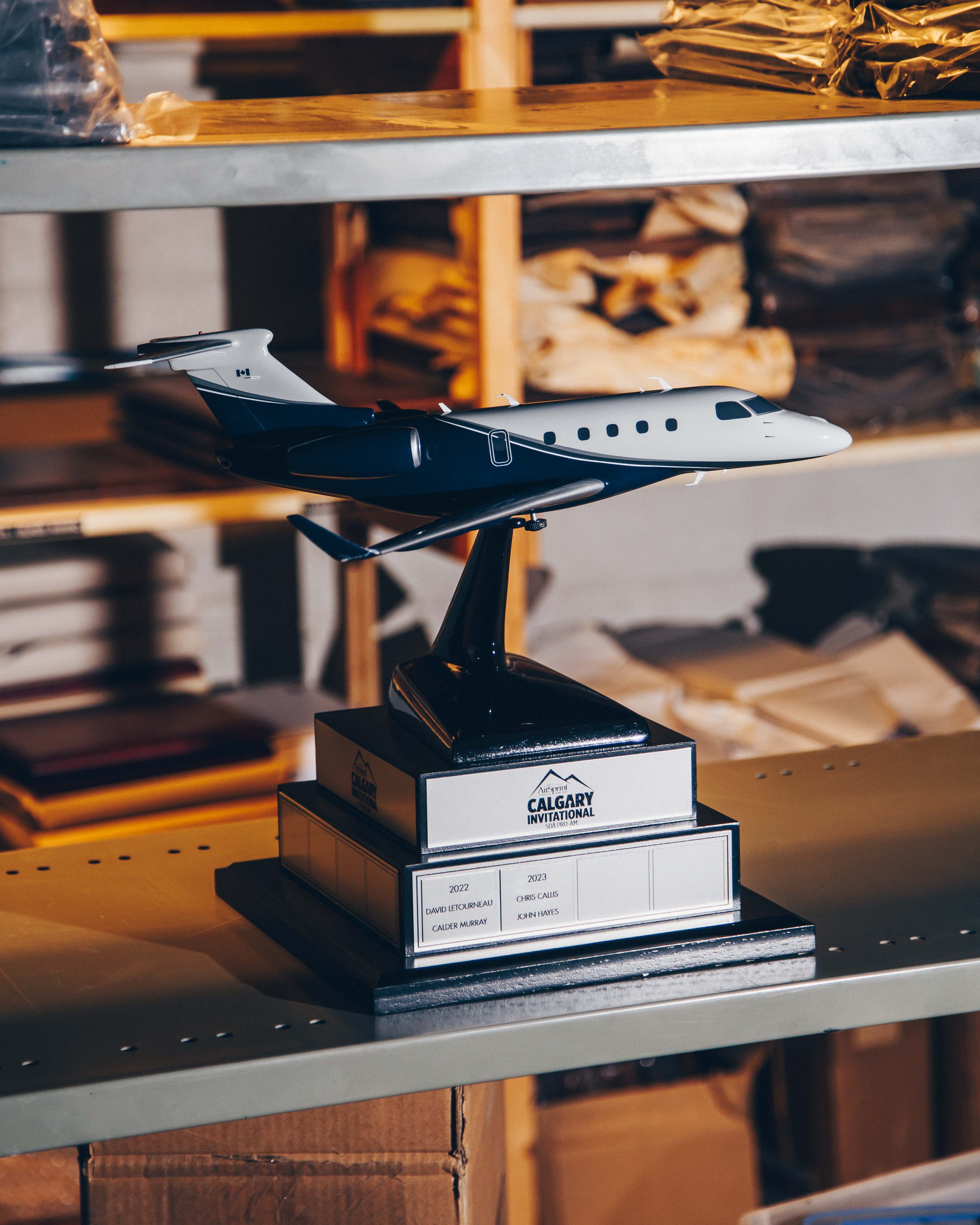 A model airplane trophy prominently displayed on a shelf in what appears to be a storage or workshop area. The trophy consists of a detailed model of a jet airplane mounted on a black pedestal with a plaque. The plaque reads "Calgary Invitational" and lists names and years, indicating it is an award. The background is filled with shelves containing various items, including boxes and fabric, suggesting an industrial or storage setting. The lighting is warm and slightly diffused, casting soft shadows and creating a cozy atmosphere. The overall mood is one of quiet achievement and industriousness.