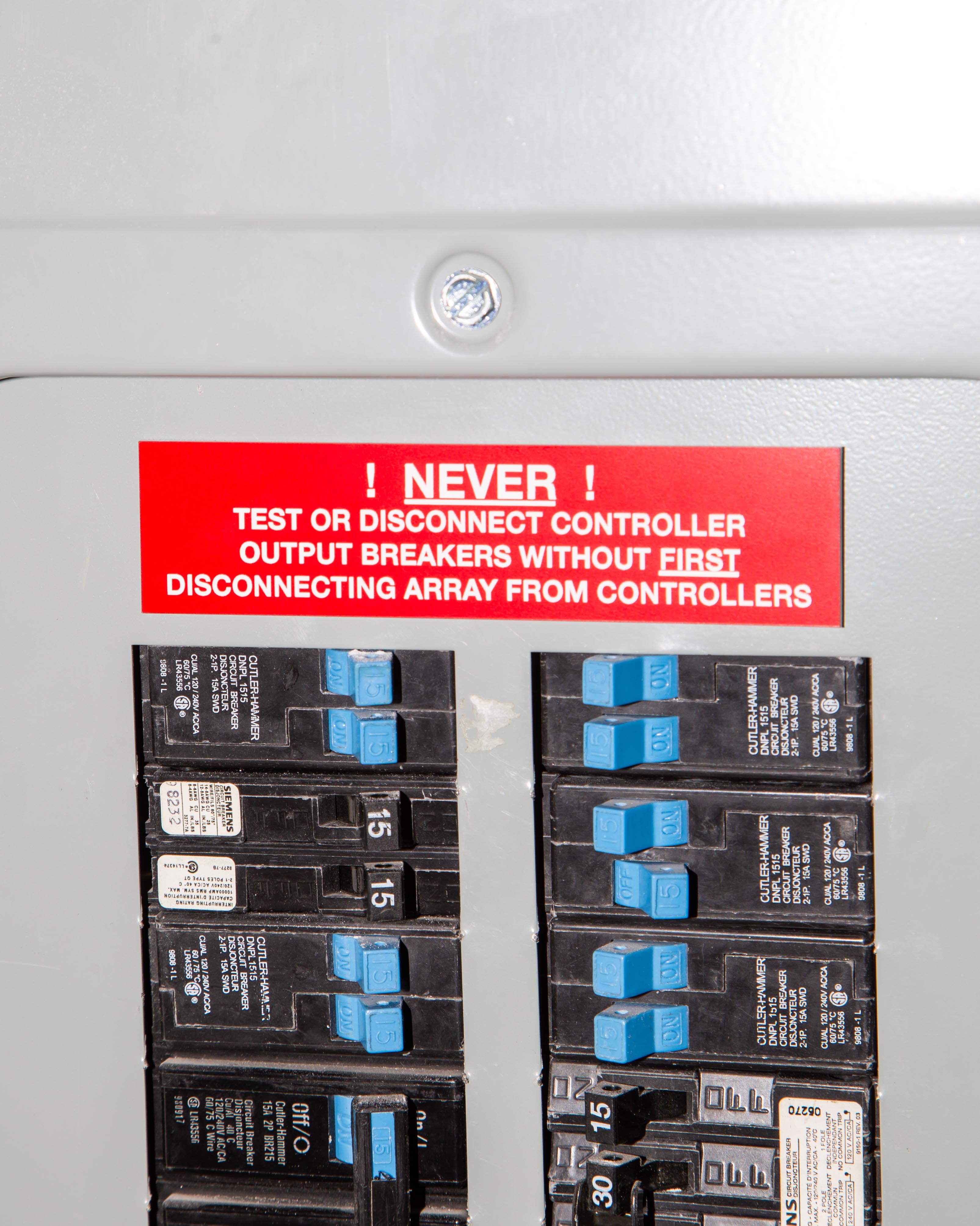 Electrical panel with a red warning sign about disconnecting controllers before testing breakers.