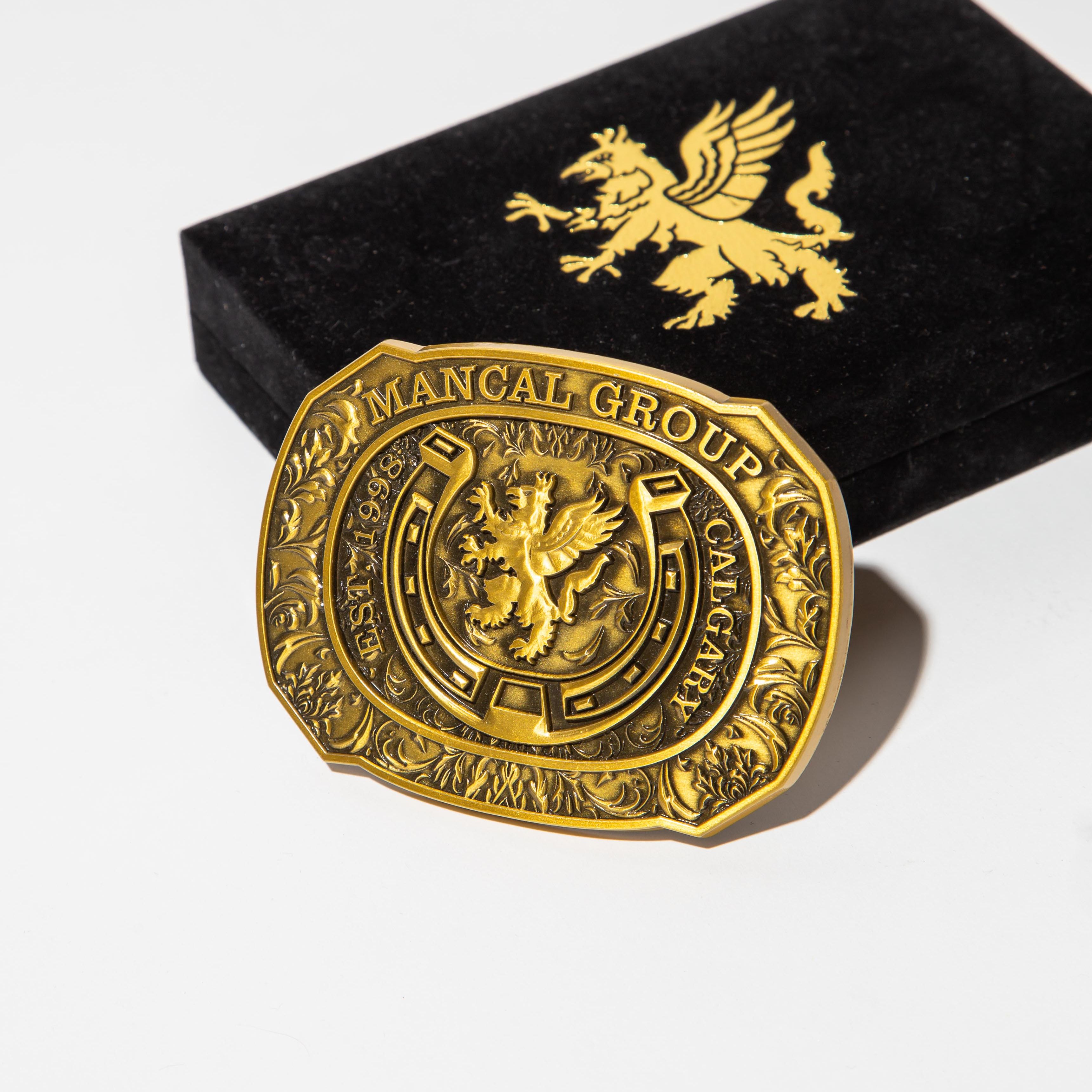 A detailed, ornate gold belt buckle with intricate engravings and a central emblem of a griffin. The buckle is prominently displayed against a plain white background, with a black velvet box partially visible behind it. The box also features a gold griffin emblem on its surface. The lighting is bright and direct, casting subtle shadows and highlighting the metallic sheen of the buckle.