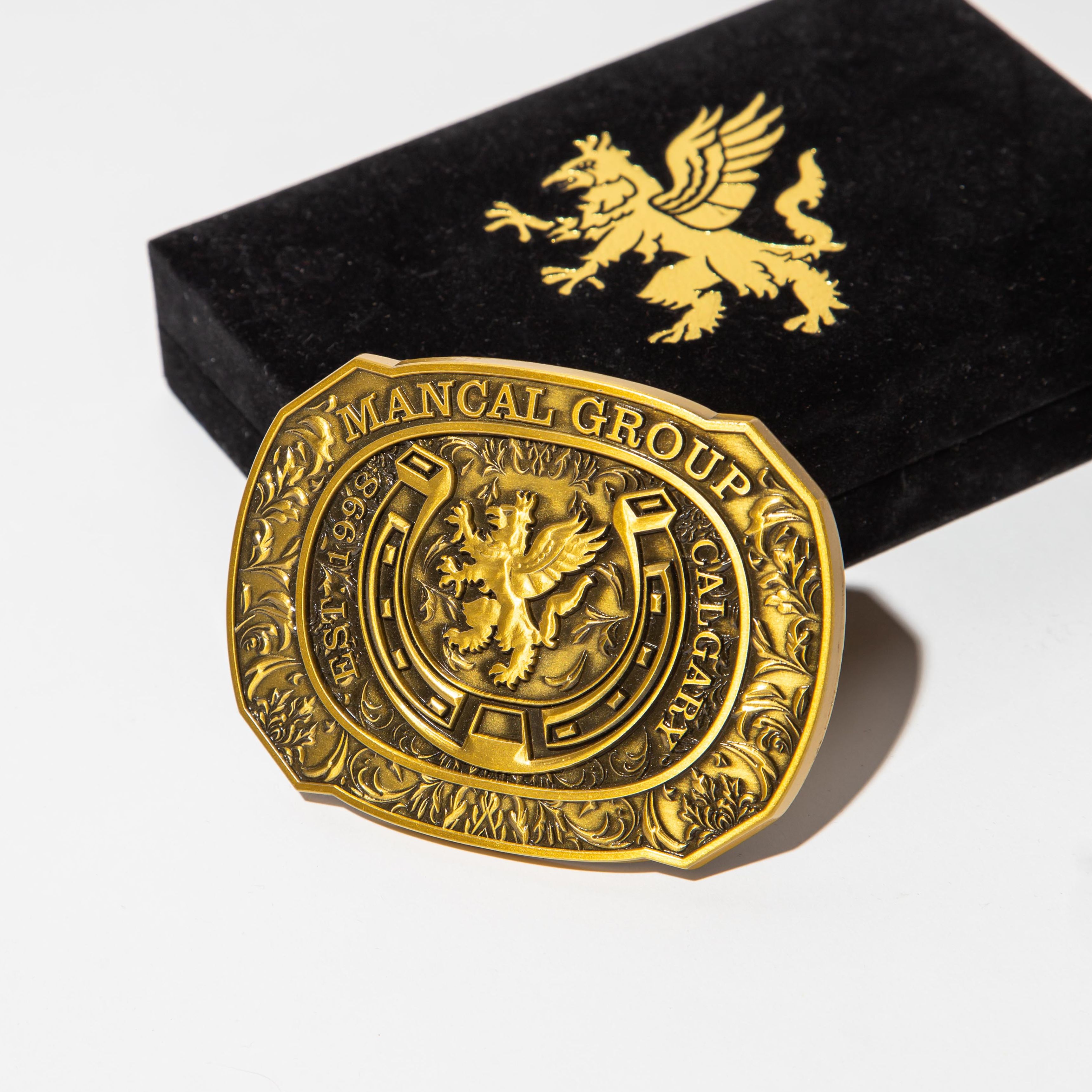 A detailed, ornate gold belt buckle with intricate engravings and a central emblem of a griffin. The buckle is prominently displayed against a plain white background, with a black velvet box partially visible behind it. The box also features a gold griffin emblem on its surface. The lighting is bright and direct, casting subtle shadows and highlighting the metallic sheen of the buckle.