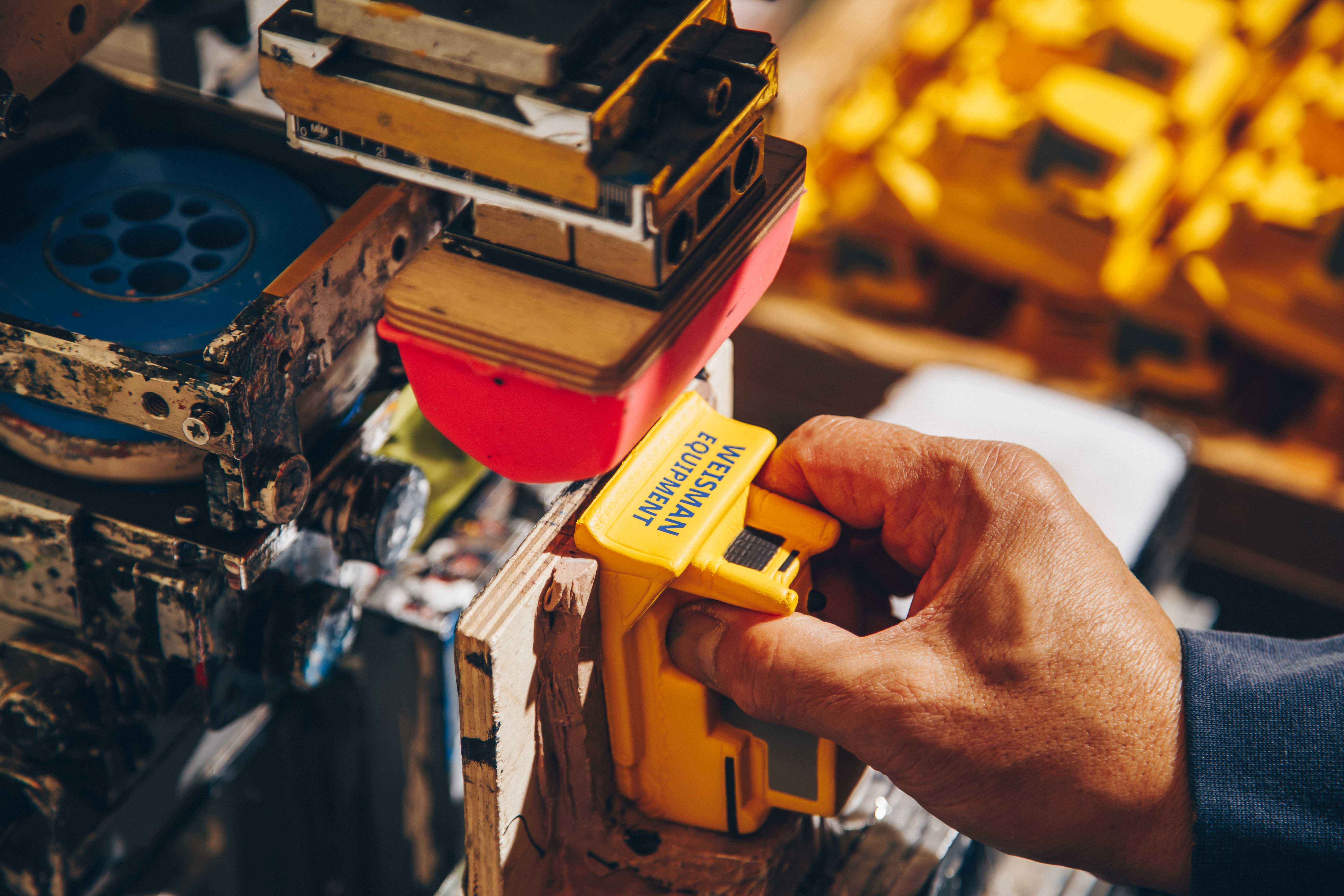 A worker uses a pad printing machine to brand a yellow plastic product labeled "Wesmann Equipment." The setup includes a red silicone transfer pad and a detailed industrial work environment for custom branding.