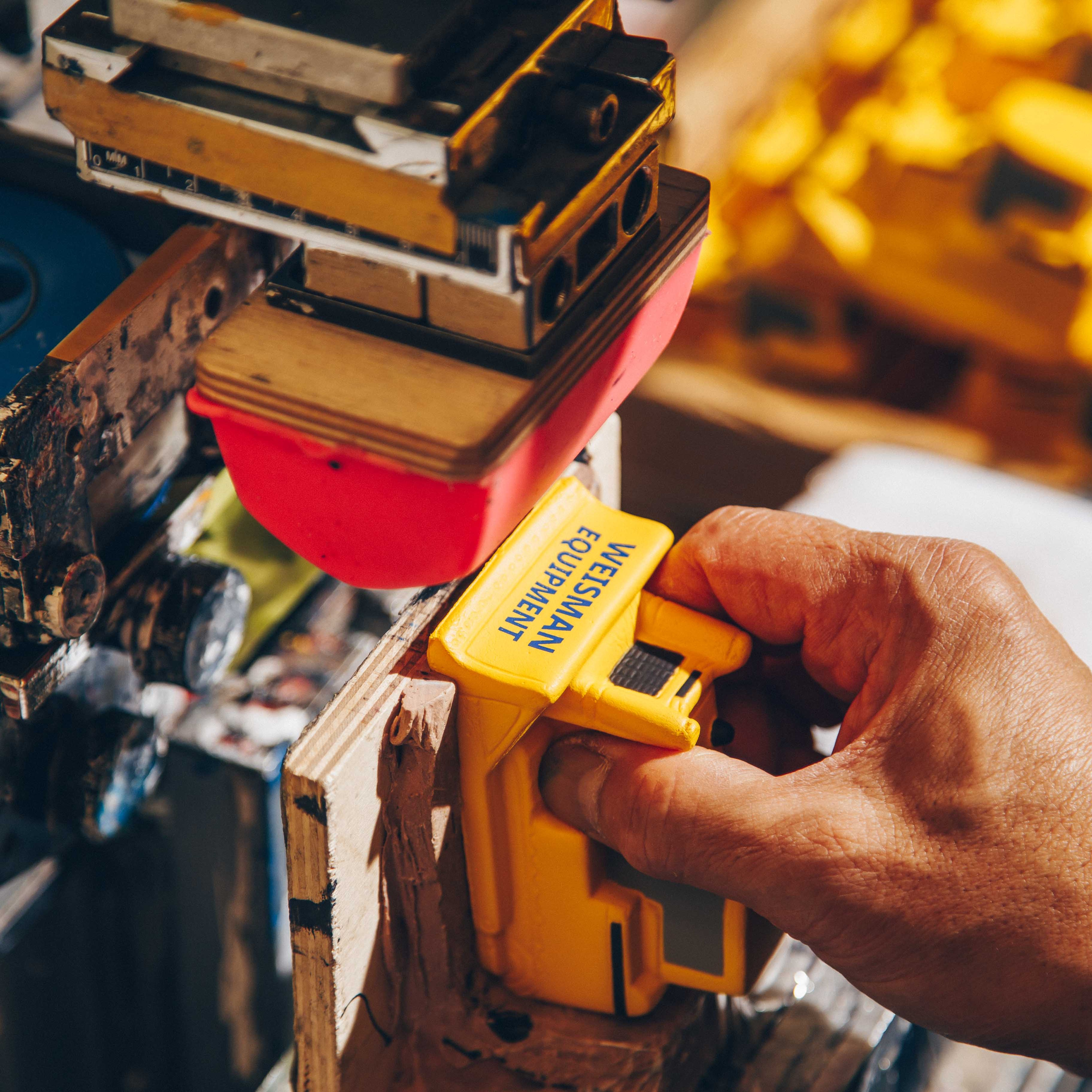 A worker uses a pad printing machine to brand a yellow plastic product labeled "Wesmann Equipment." The setup includes a red silicone transfer pad and a detailed industrial work environment for custom branding.