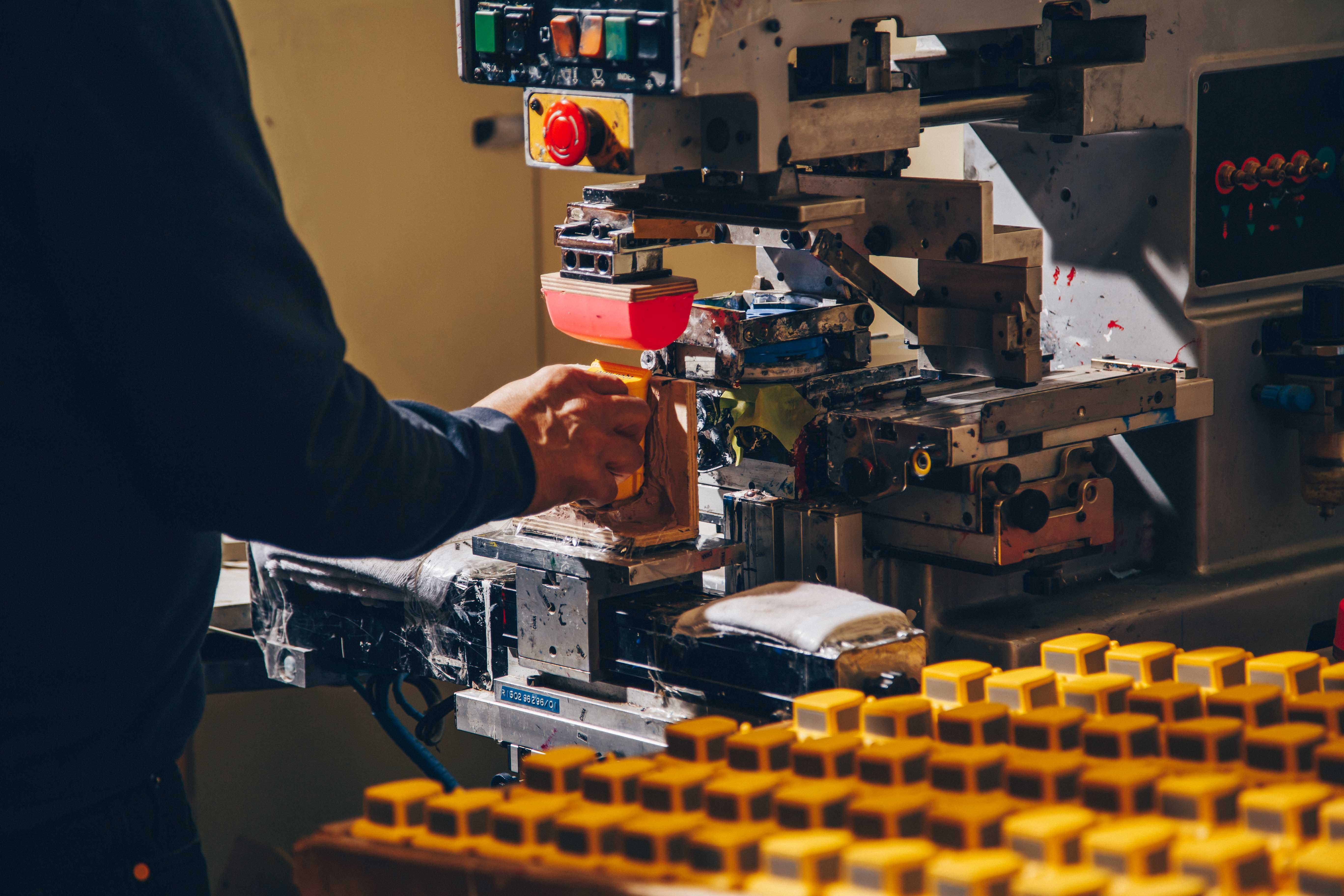 A worker operating a pad printing machine to brand yellow plastic components. The machine features a red silicone transfer pad, with a tray of identical yellow parts visible in the foreground. The setup showcases precision and efficiency in industrial product branding and customization.
