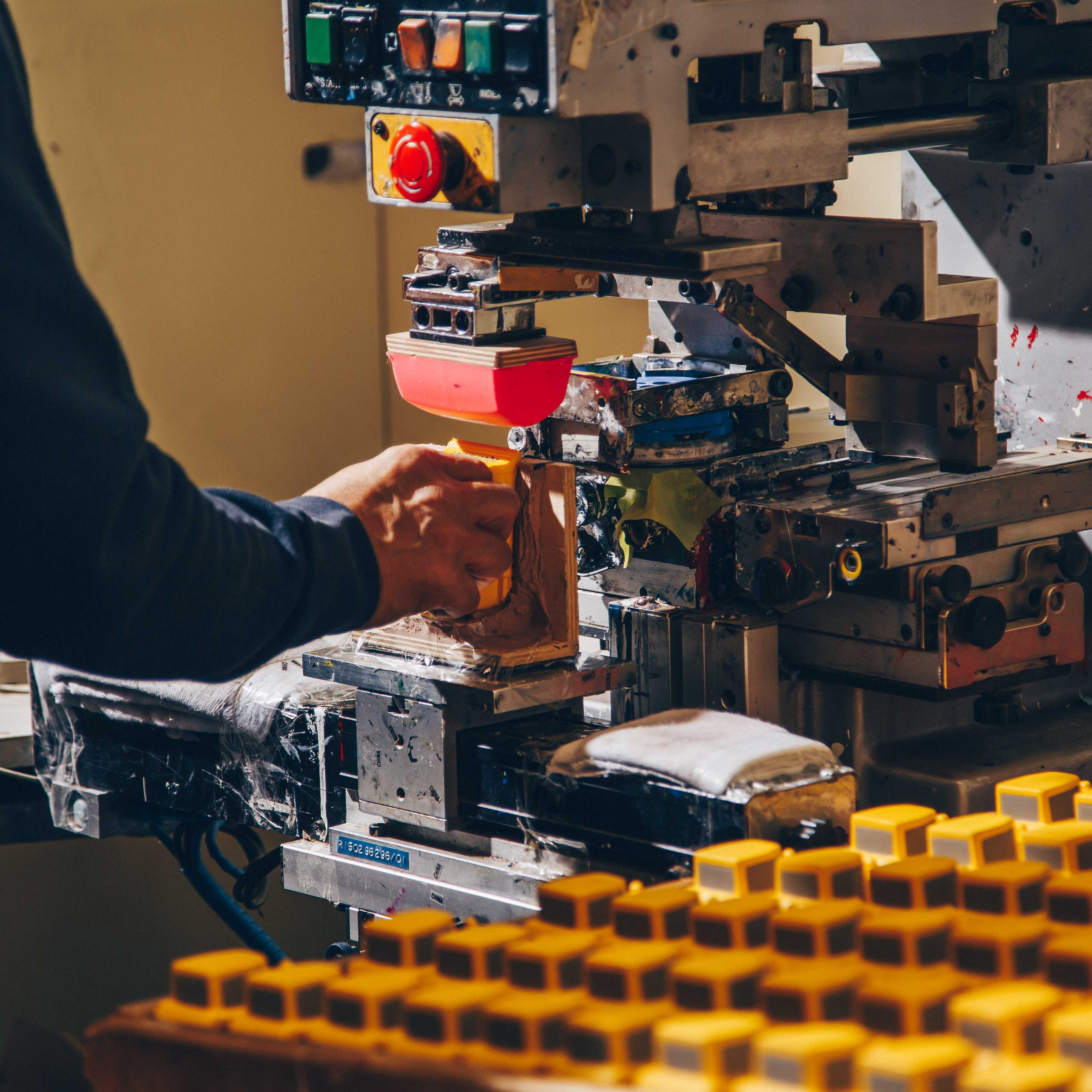 A worker operating a pad printing machine to brand yellow plastic components. The machine features a red silicone transfer pad, with a tray of identical yellow parts visible in the foreground. The setup showcases precision and efficiency in industrial product branding and customization.