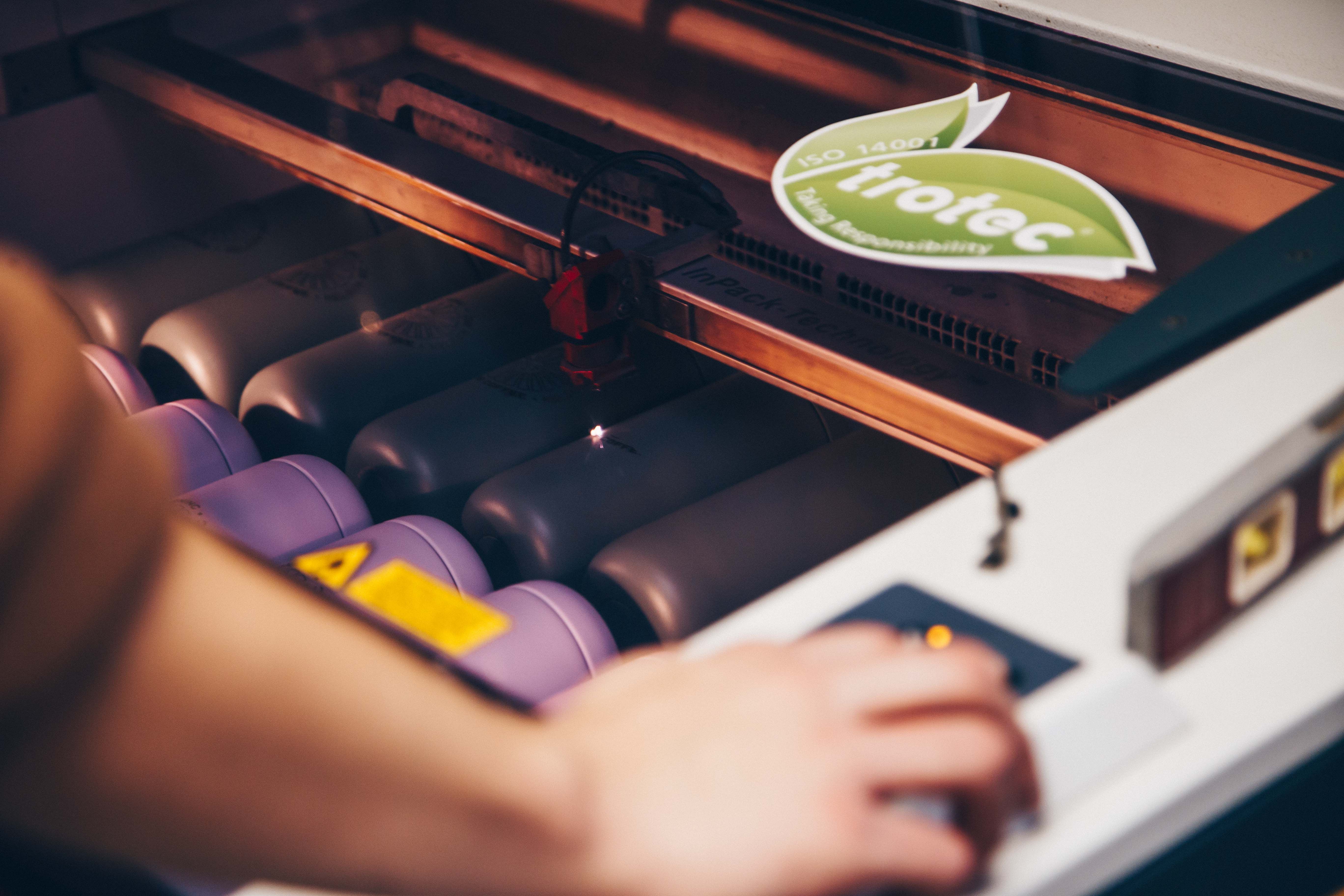 A laser engraving machine etching designs onto rows of drinkware. The machine’s operator adjusts settings, ensuring precision in the branding process. A green sustainability sticker is visible, emphasizing eco-conscious production.