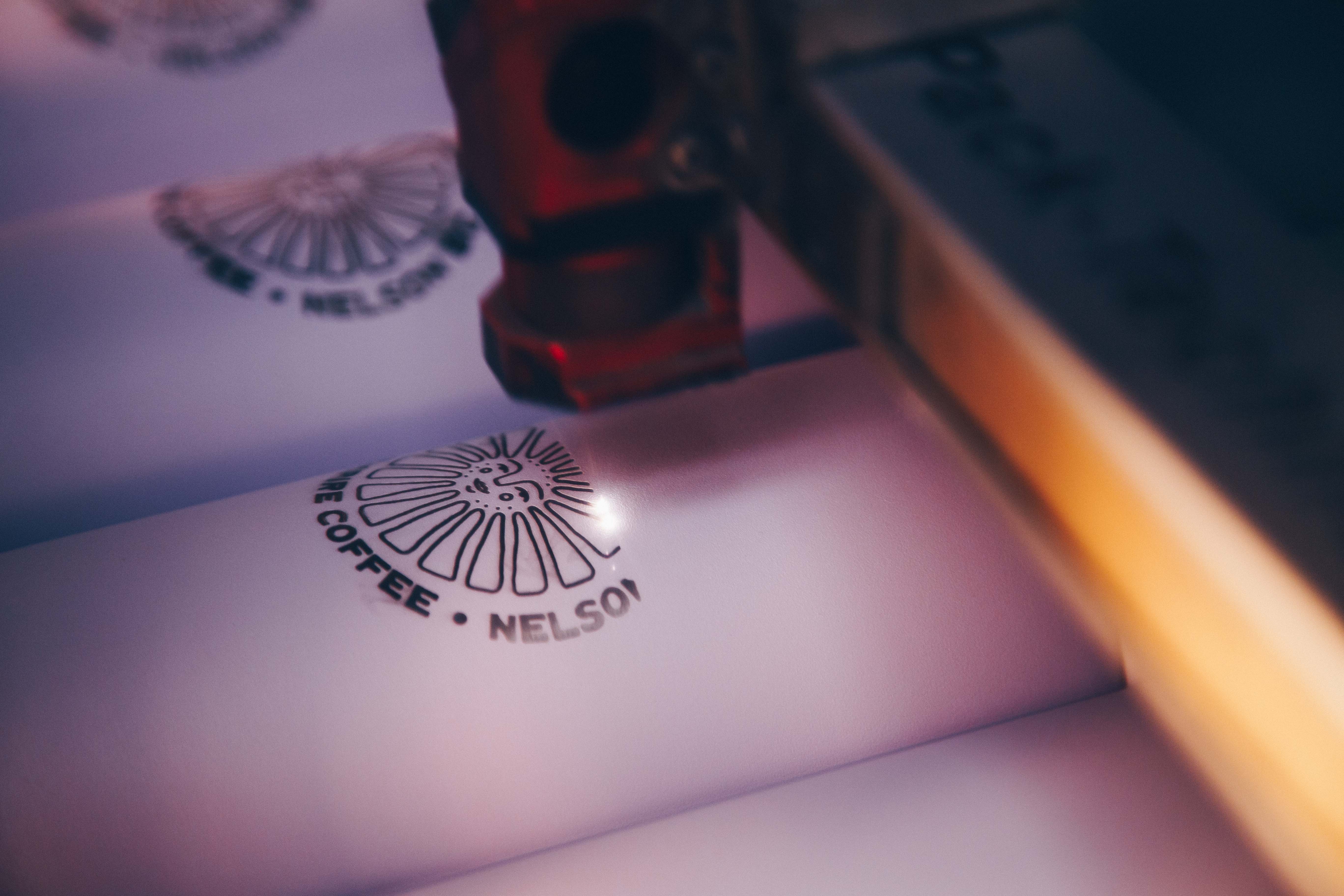 A close-up of a laser engraving process on white drinkware. The design features the logo "Good Morning Coffee, Nelson BC" being precisely engraved, showcasing customization for branded tumblers and mugs.