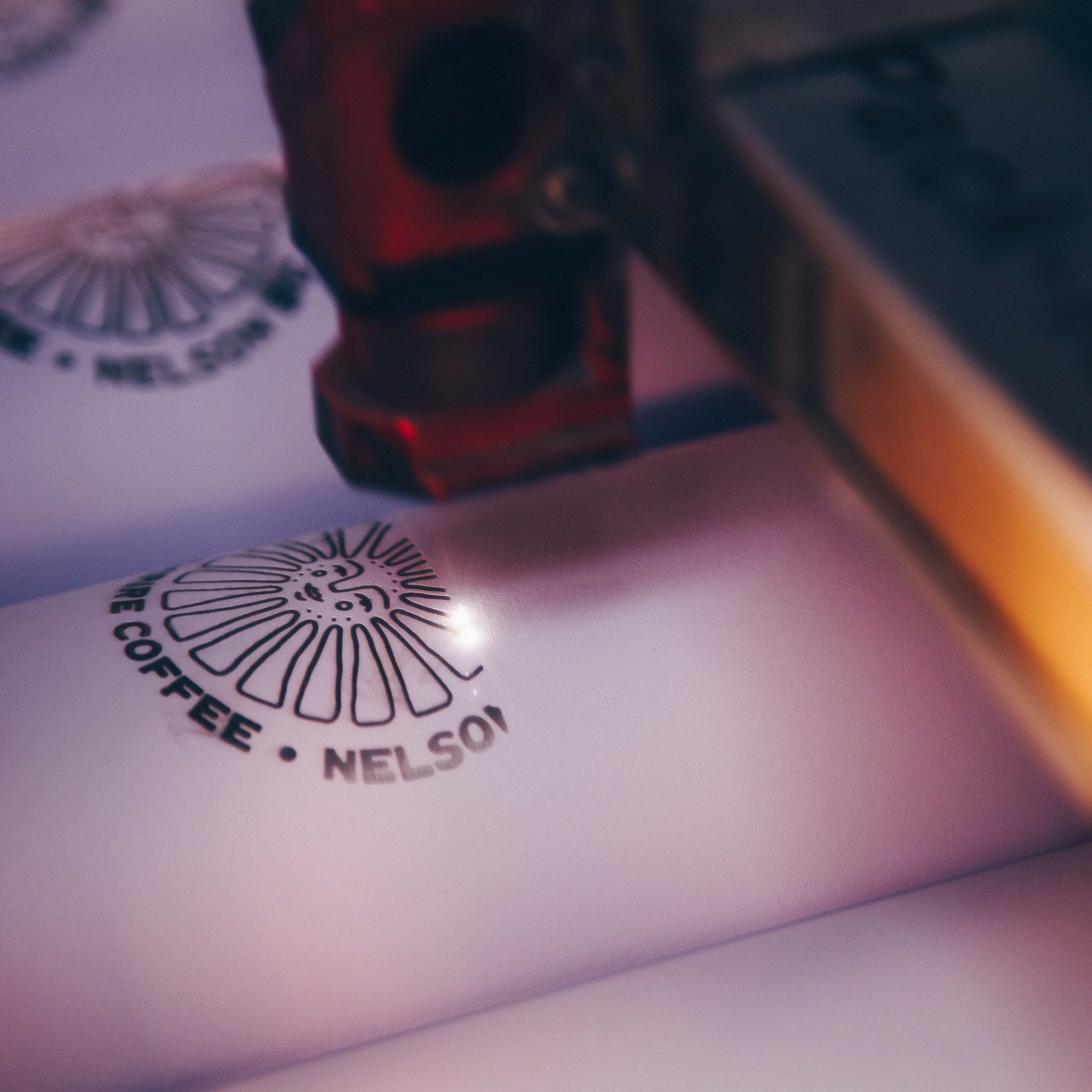A close-up of a laser engraving process on white drinkware. The design features the logo "Good Morning Coffee, Nelson BC" being precisely engraved, showcasing customization for branded tumblers and mugs.