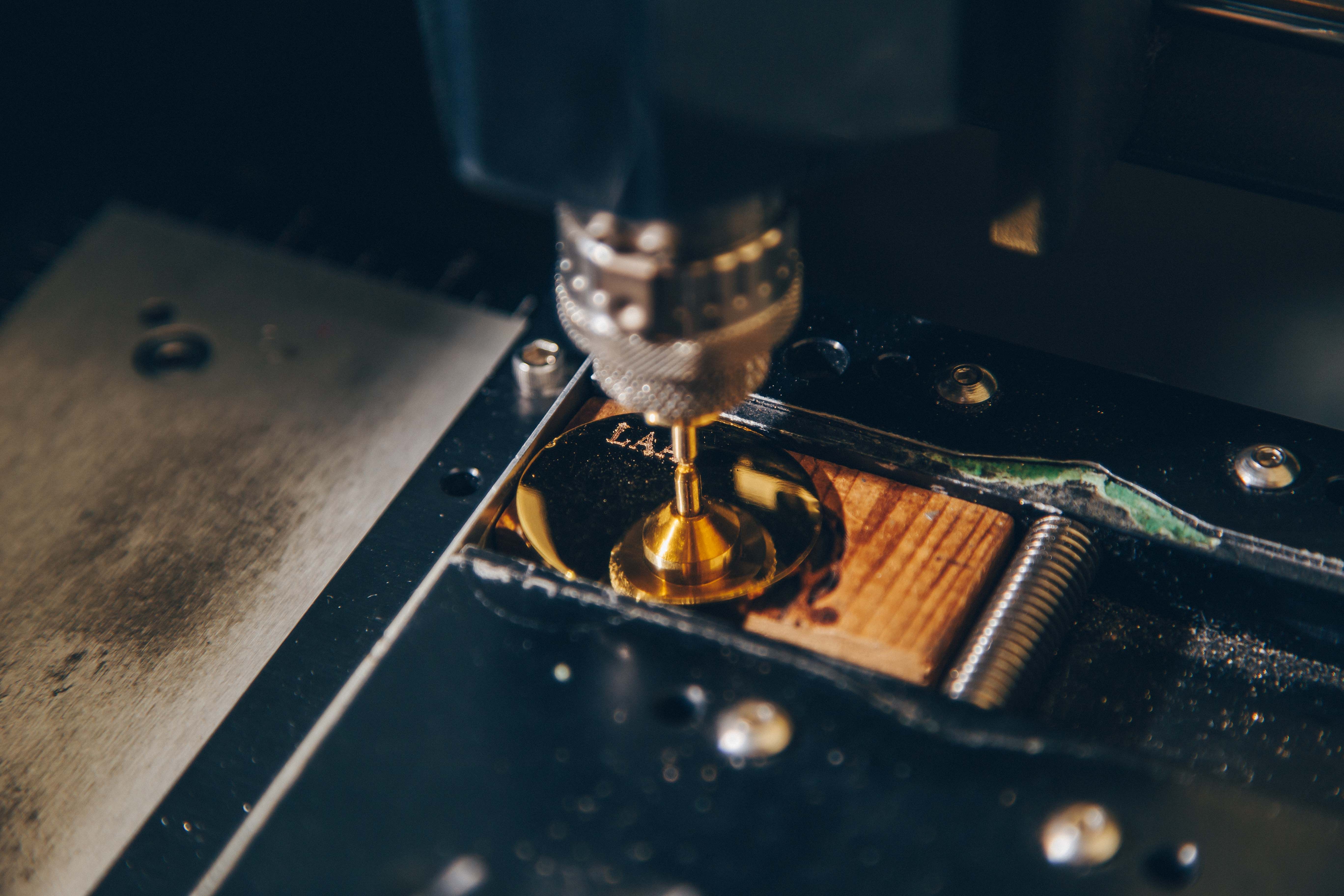 A close-up view of a CNC machine in operation. The focus is on the drill bit, which is actively engaged in machining a piece of material. The material appears to be a combination of metal and wood, with the metal part being polished and reflective, possibly brass or gold-colored. The drill bit is metallic and has a textured grip, indicating precision engineering. The surrounding area includes parts of the machine, such as screws and a metal plate, which are slightly out of focus, emphasizing the drill bit and the workpiece.