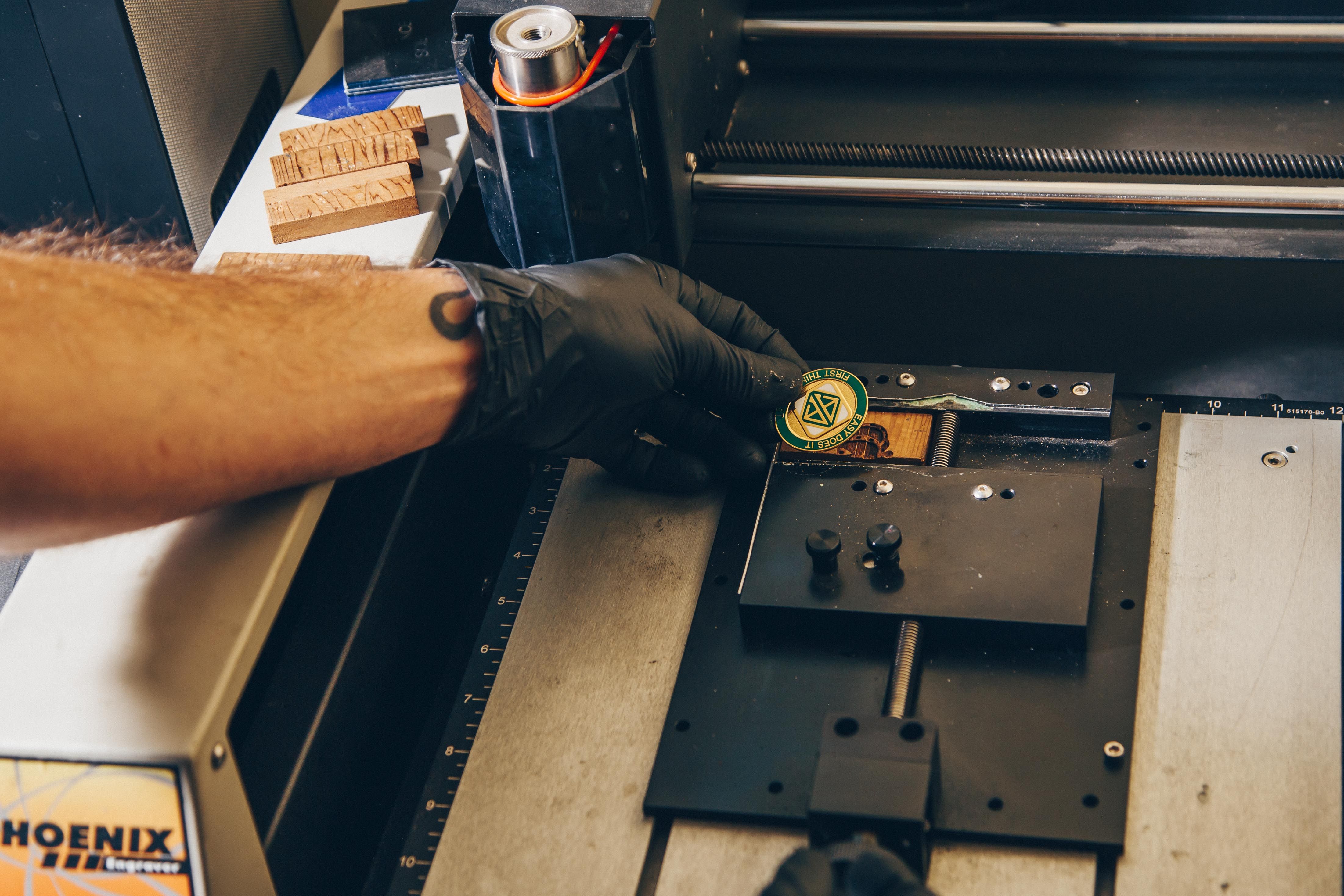 A close-up view of a person's arm, wearing a black glove, interacting with a piece of machinery. The arm has a tattoo visible on the wrist. The person is holding a circular object with a green and yellow design, possibly a badge or token, and placing it onto a metal platform. The machinery appears to be some sort of engraving or cutting device, with a ruler and various knobs and screws visible.