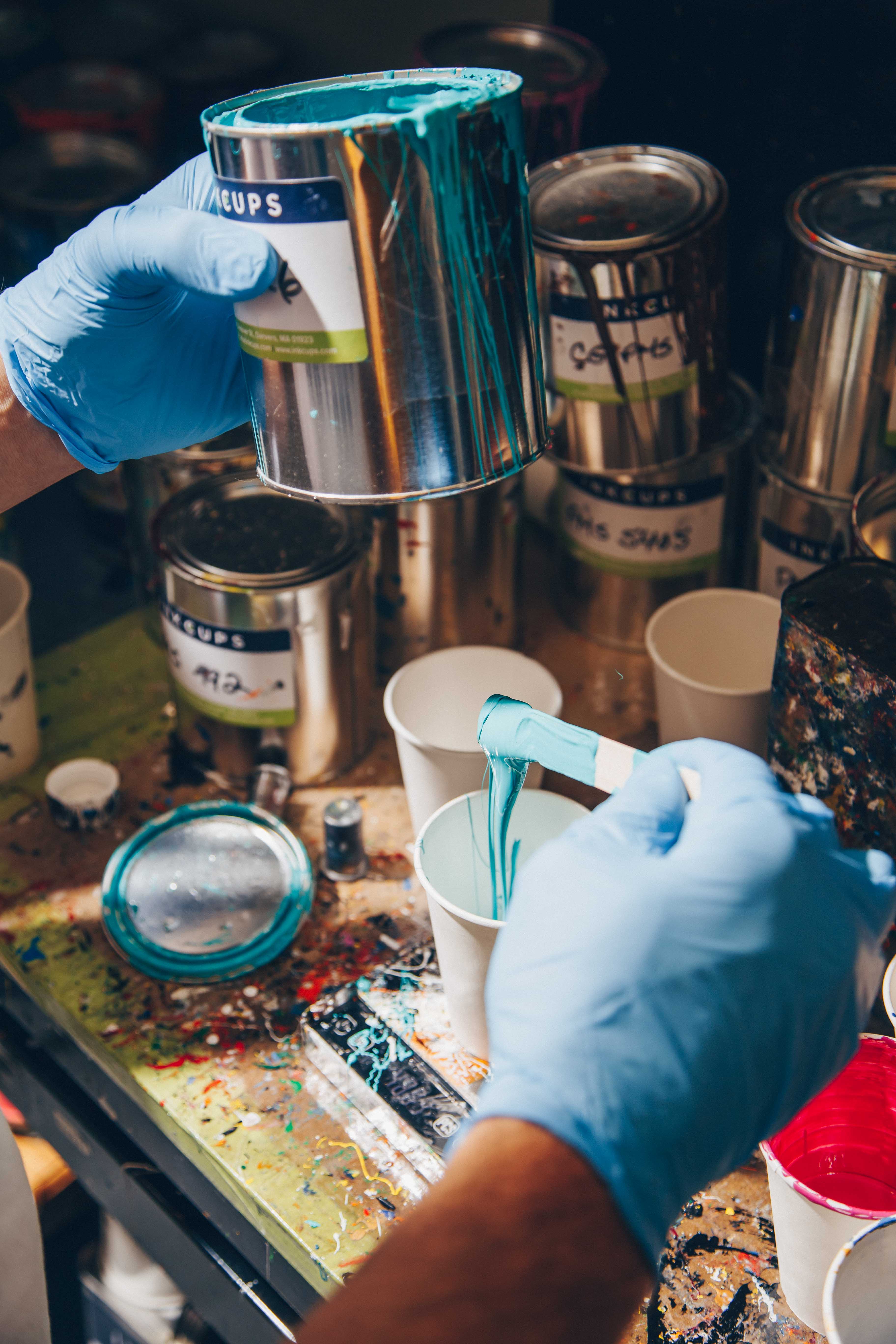 A pad printing workstation with a worker in blue gloves pouring custom-mixed turquoise ink into a white cup. The desk is covered with colorful ink-filled cups and tools used for preparing pad printing inks for precision branding.