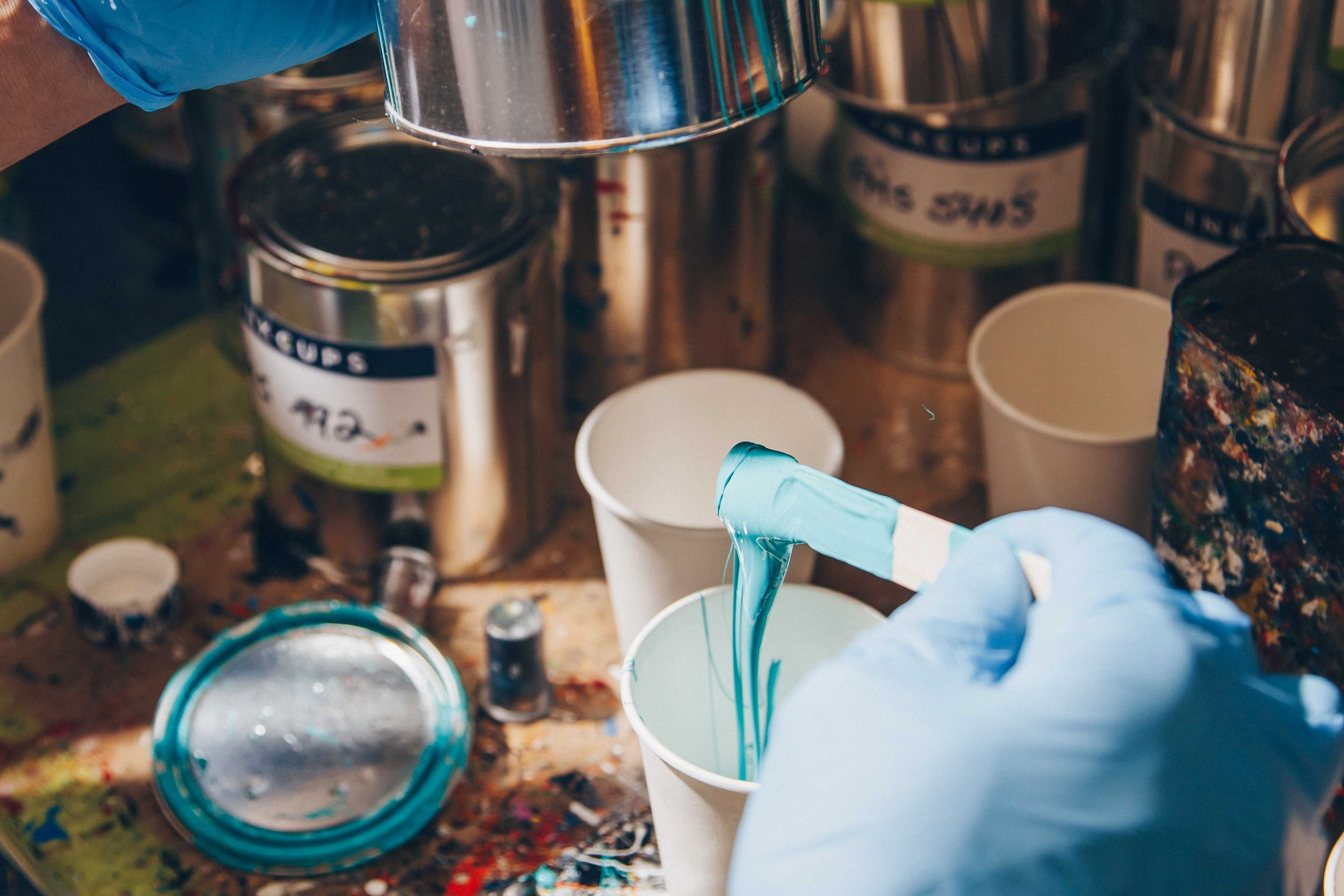 A pad printing workstation with a worker in blue gloves pouring custom-mixed turquoise ink into a white cup. The desk is covered with colorful ink-filled cups and tools used for preparing pad printing inks for precision branding.