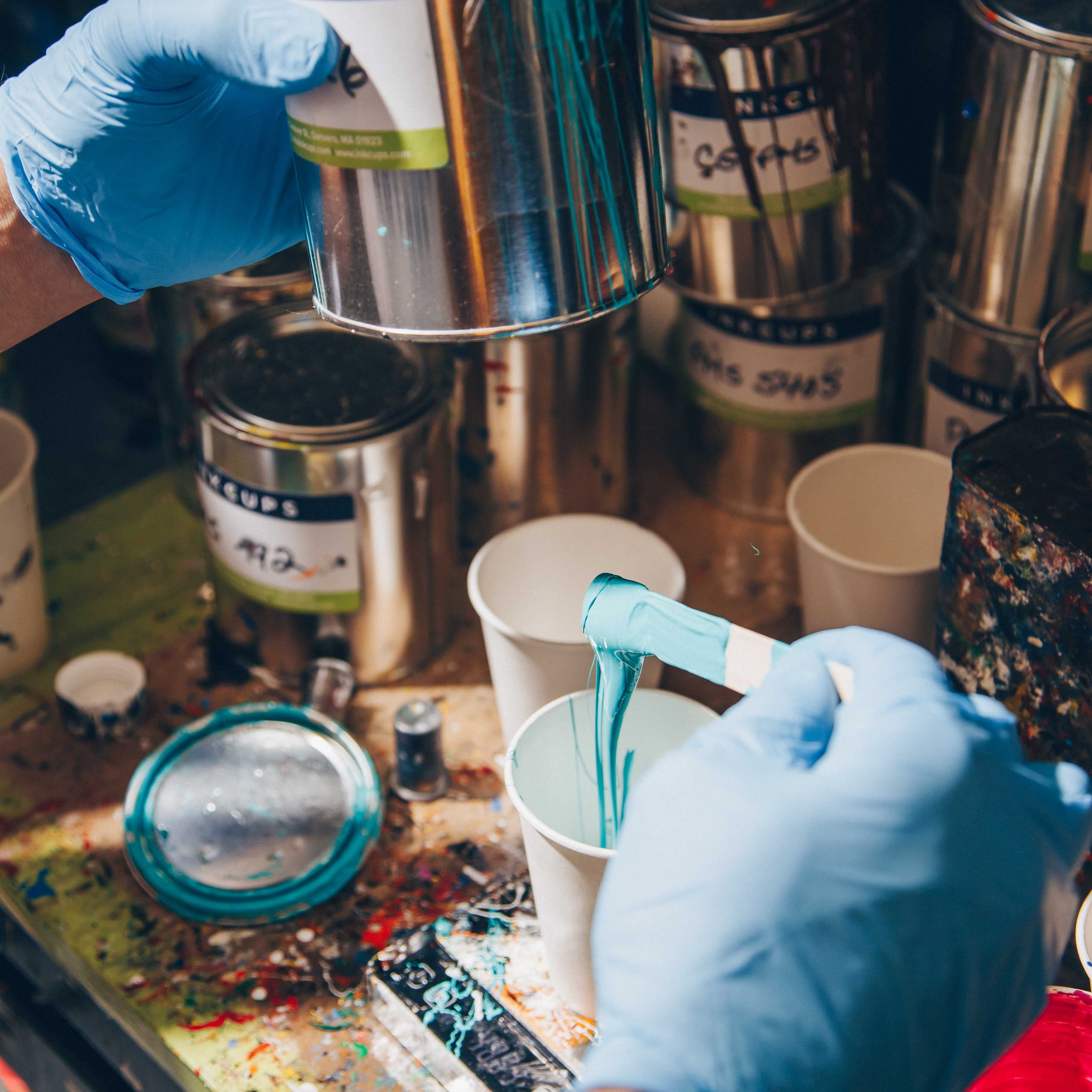 A pad printing workstation with a worker in blue gloves pouring custom-mixed turquoise ink into a white cup. The desk is covered with colorful ink-filled cups and tools used for preparing pad printing inks for precision branding.